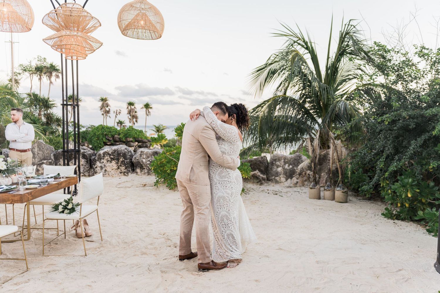 Couple shares first dance on sandy beach at tropical wedding venue with palm trees and boho decor at sunset.