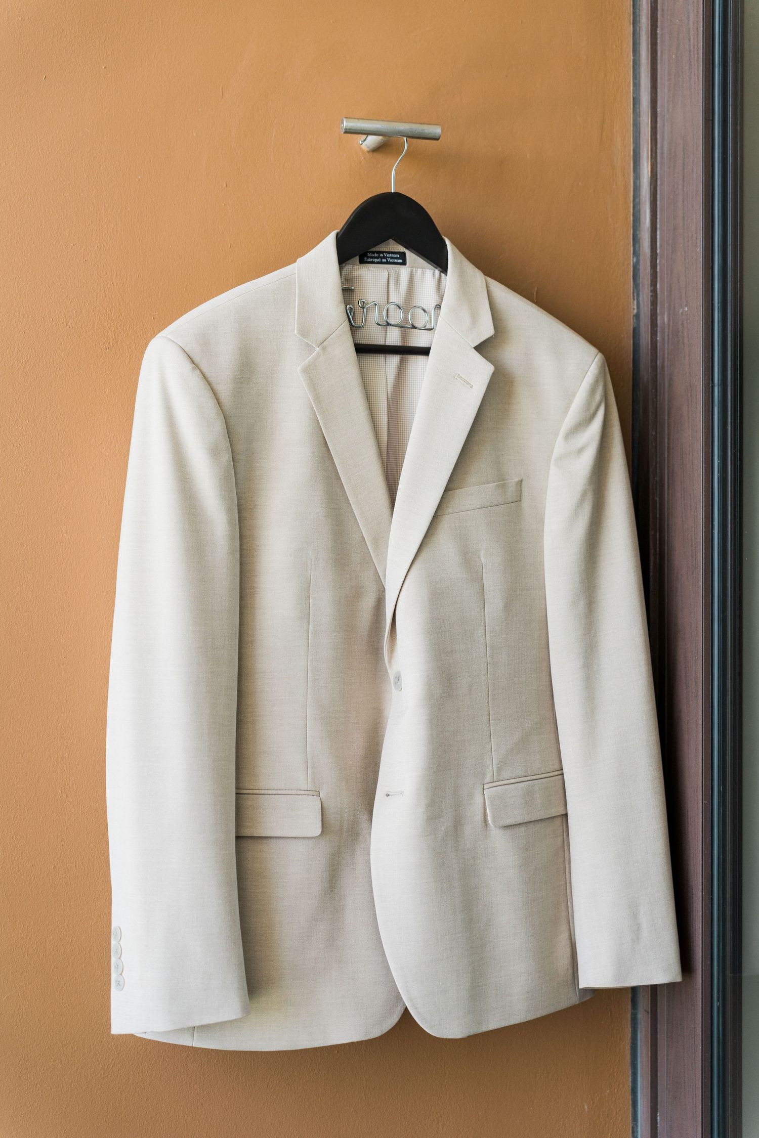 Xcaret Arte Wedding - Cream white men's blazer jacket hanging on a black hanger against a warm beige wall.