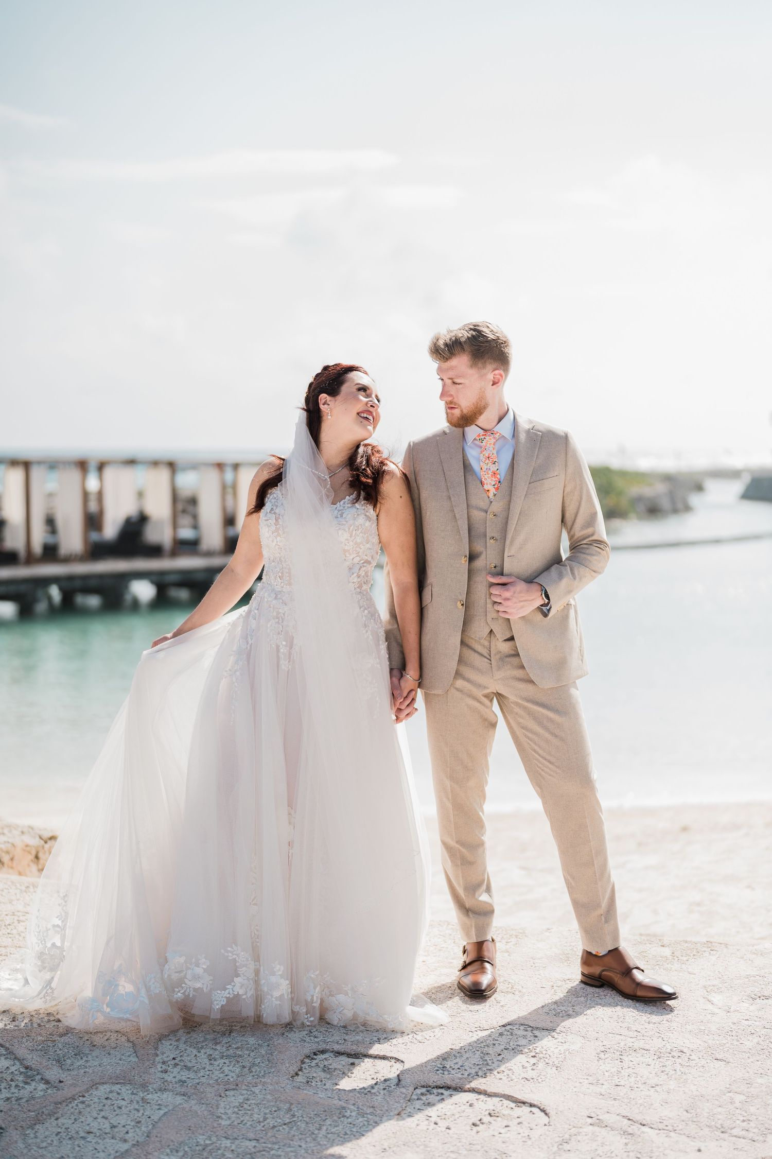Bride and groom pose romantically by the water on their wedding day under a bright blue sky.