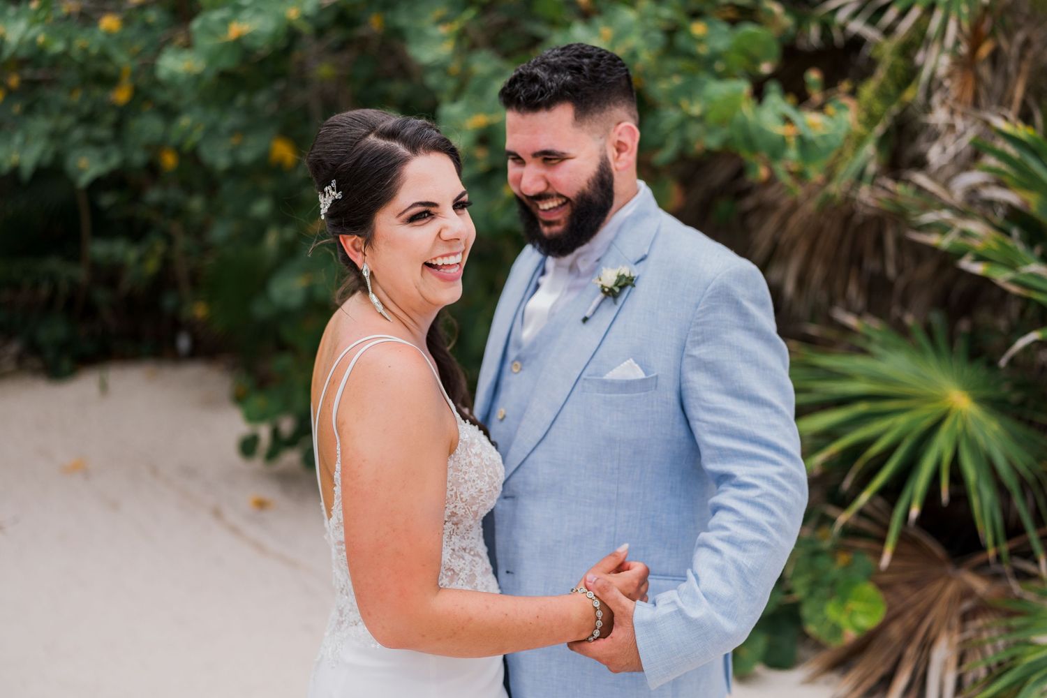 Smiling bride and bearded groom in light blue suit share a joyful moment on a tropical beach.