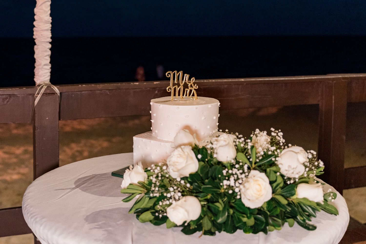 Elegant two-tier white wedding cake with Mr and Mrs topper surrounded by white roses on a table.