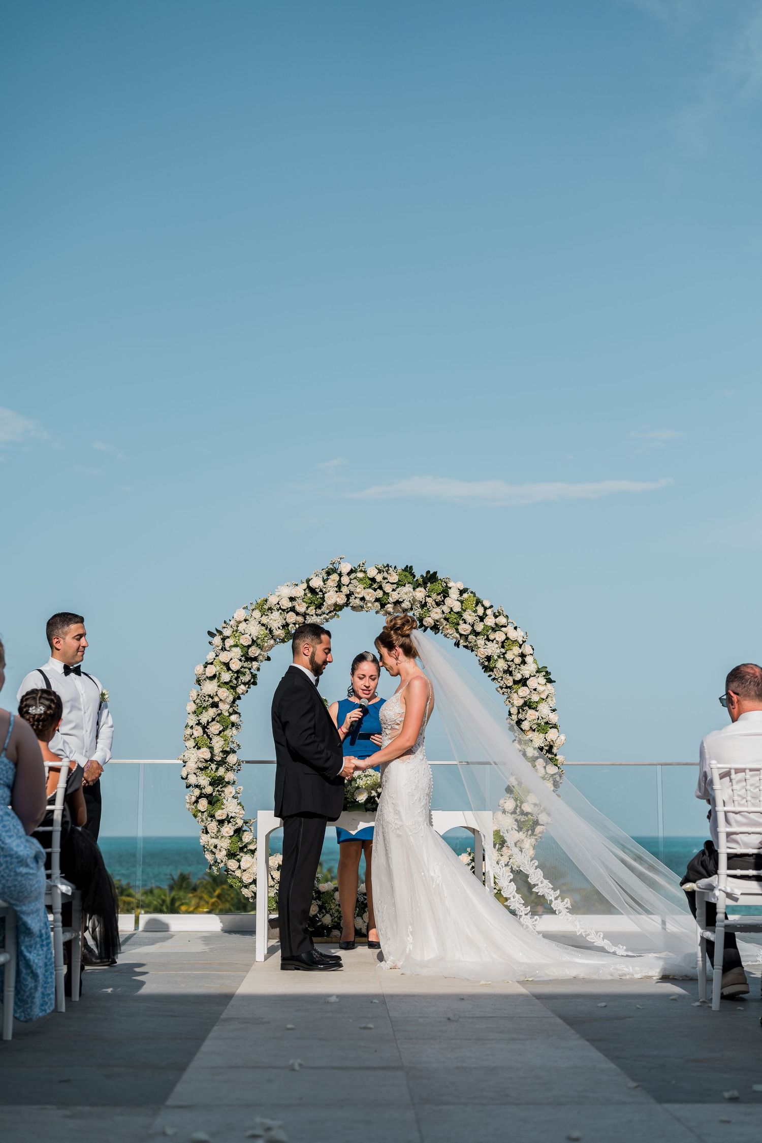 Bride and groom holding hands at altar under circular floral arch during oceanfront wedding ceremony.