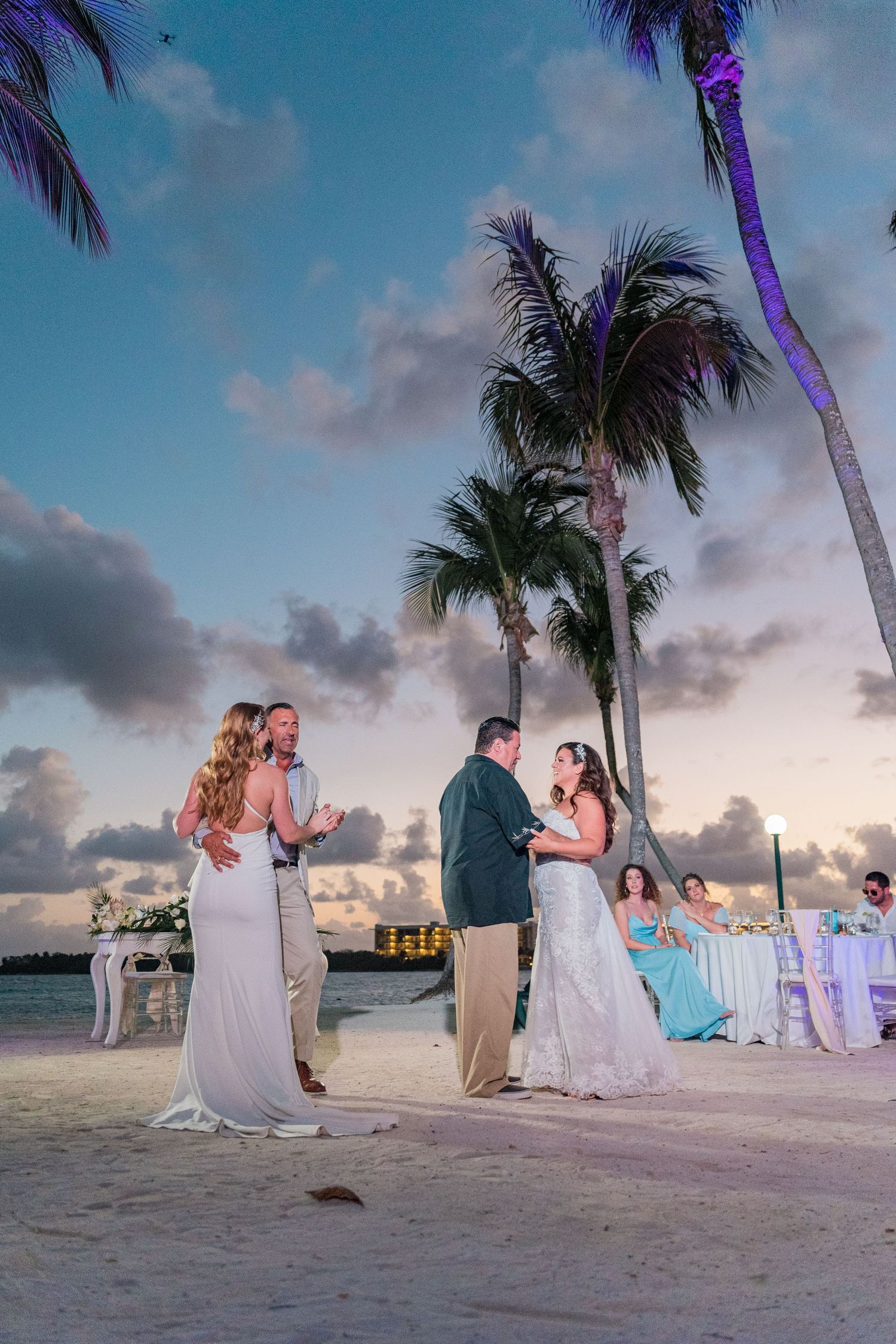 Two brides dancing with partners on beach at sunset with palm trees and guests.