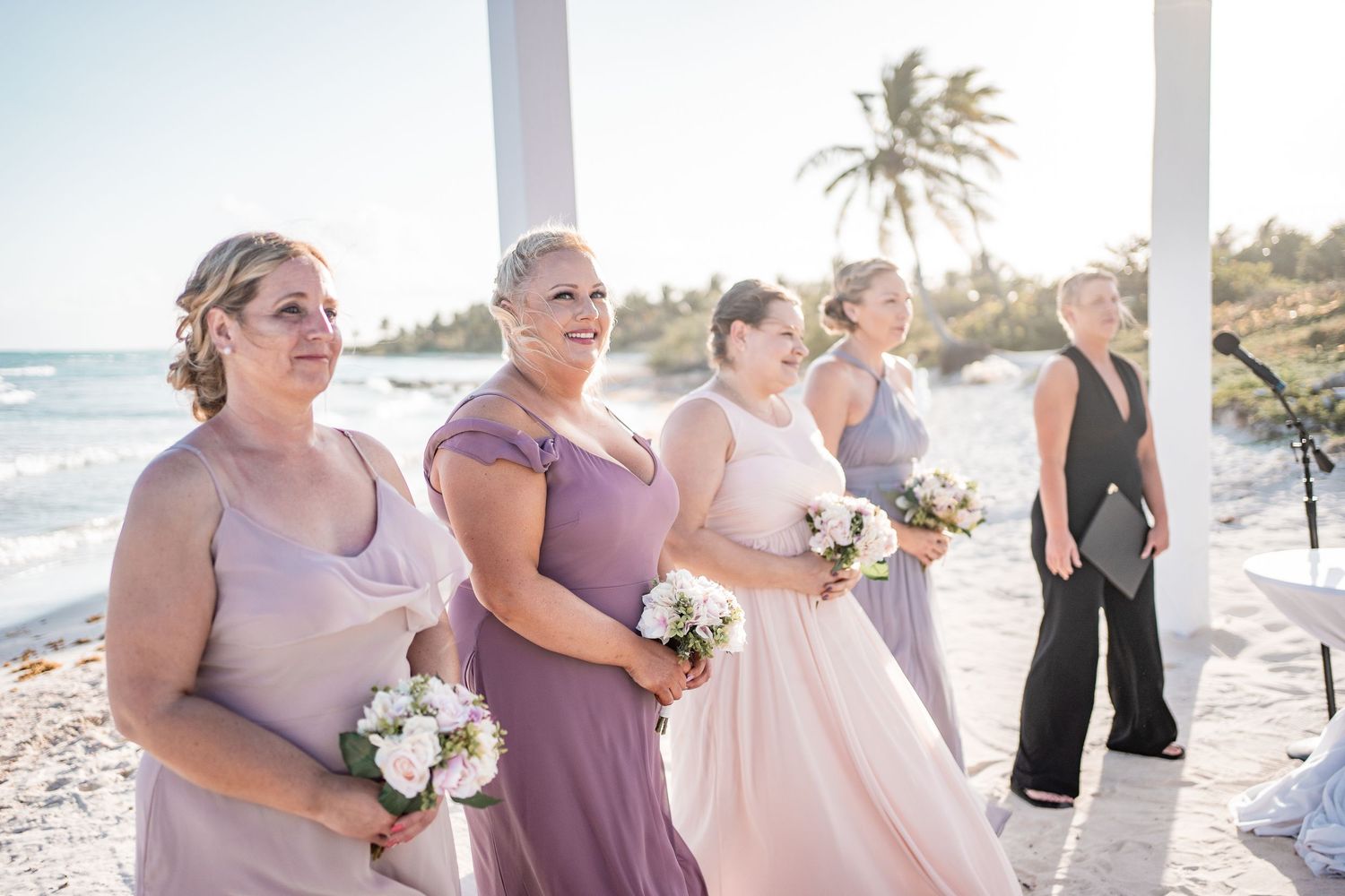 Bridesmaids in lavender dresses holding white bouquets stand at a beach wedding ceremony with ocean views.