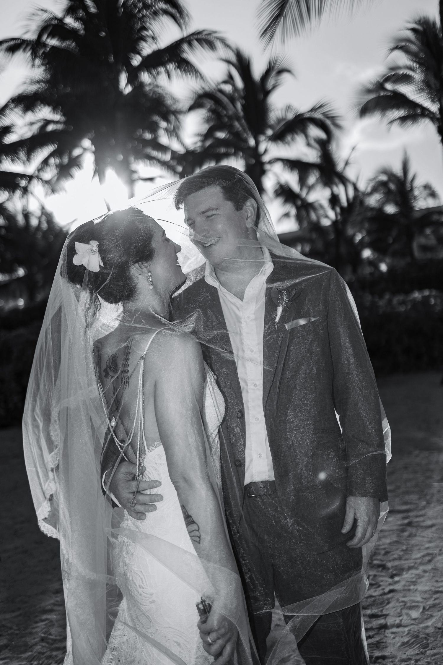 Black and white photo of a bride and groom sharing a romantic moment under a veil with tropical palm trees at sunset.