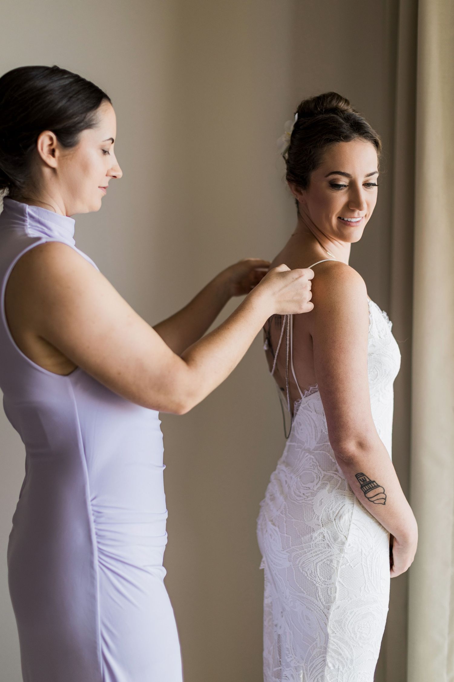 Bridesmaid in lavender dress helping bride in white lace gown with back buttons before wedding ceremony.
