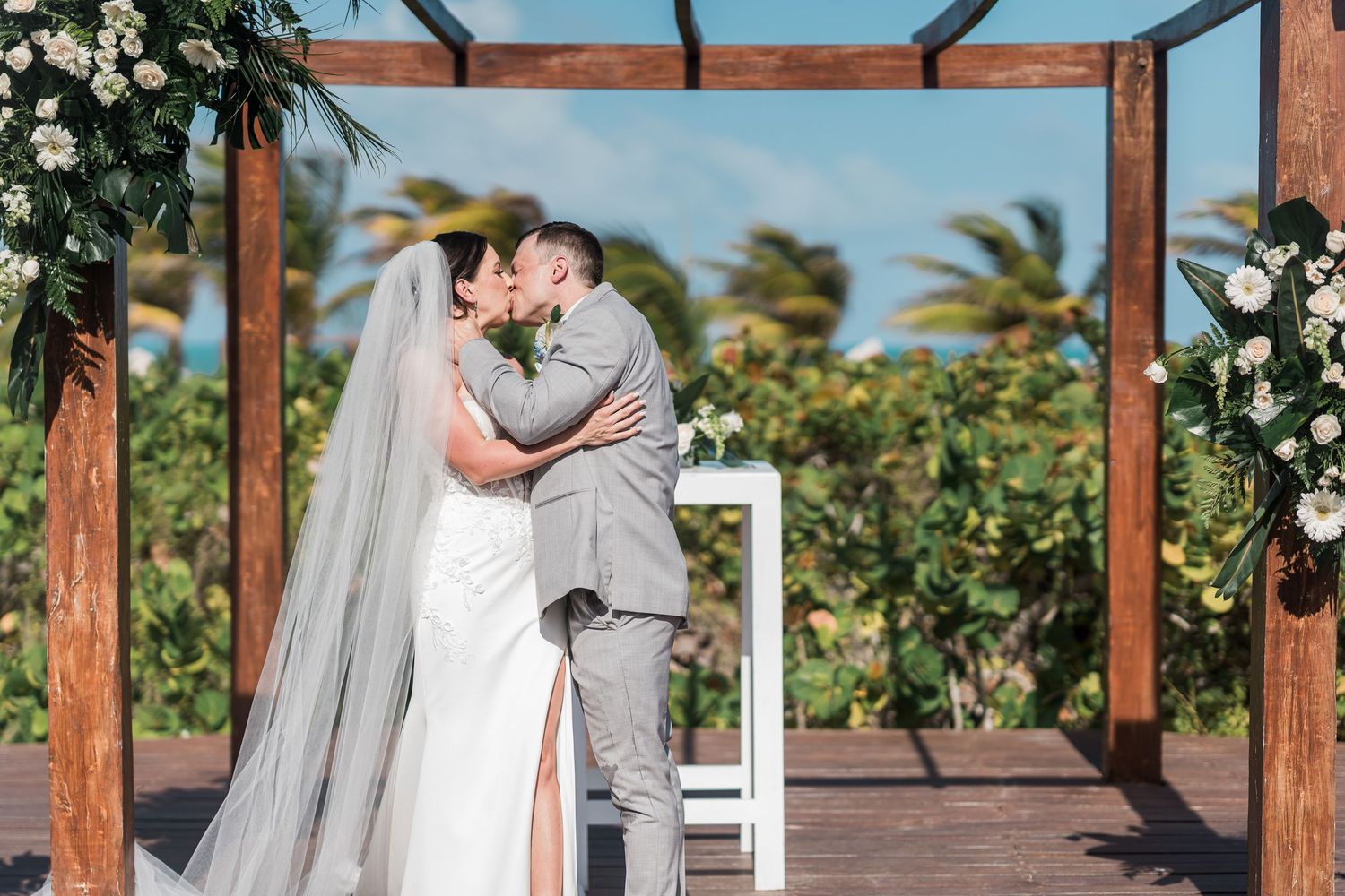 Bride and groom share first kiss under wooden arch with tropical flowers at outdoor beach wedding ceremony.
