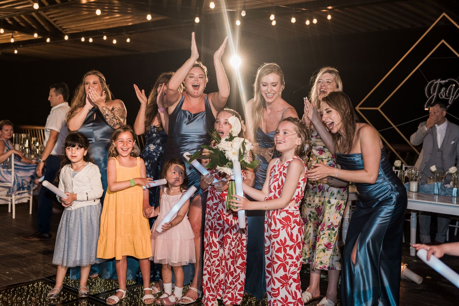 Group of women and girls posing joyfully at an elegant wedding reception, with bouquet toss and festive decorations.