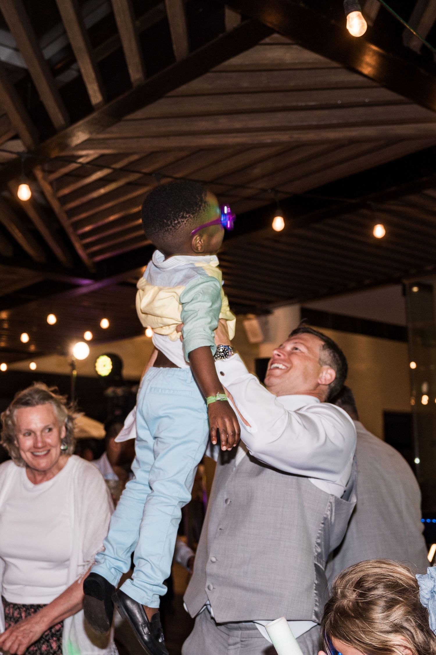 Man in gray suit joyfully lifts a young child at a wedding reception with string lights overhead.