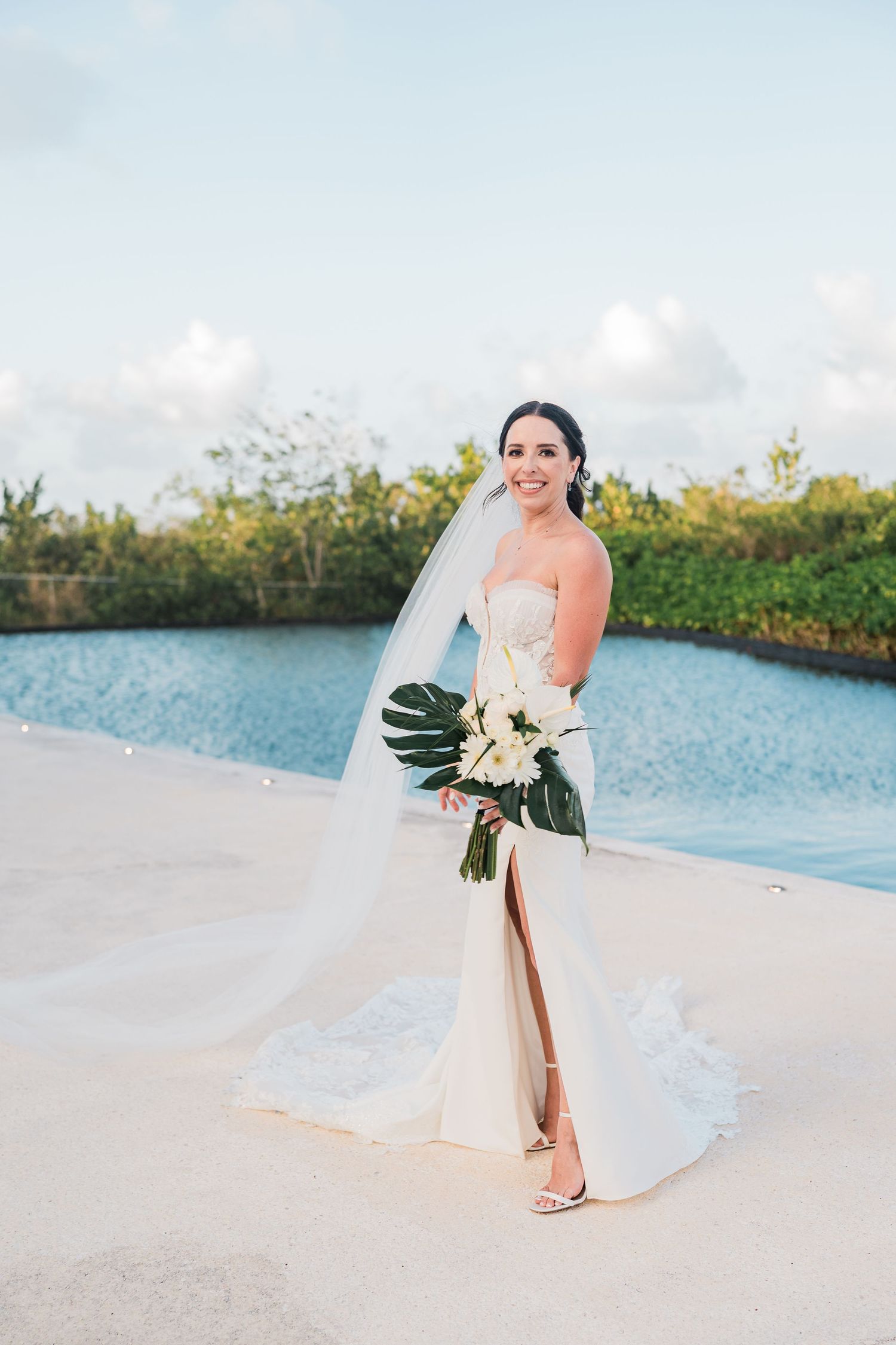Bride in strapless white gown with veil holds tropical bouquet poolside at outdoor wedding venue.