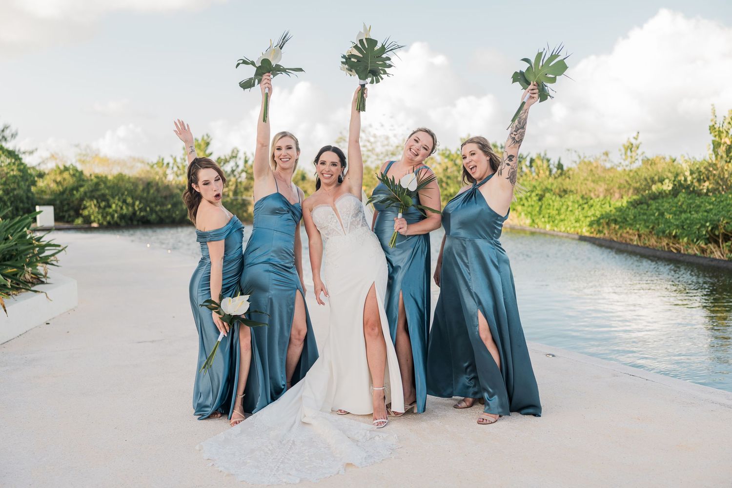 Bride in white lace gown poses with four bridesmaids in teal satin dresses on a tropical beach, arms raised joyfully.
