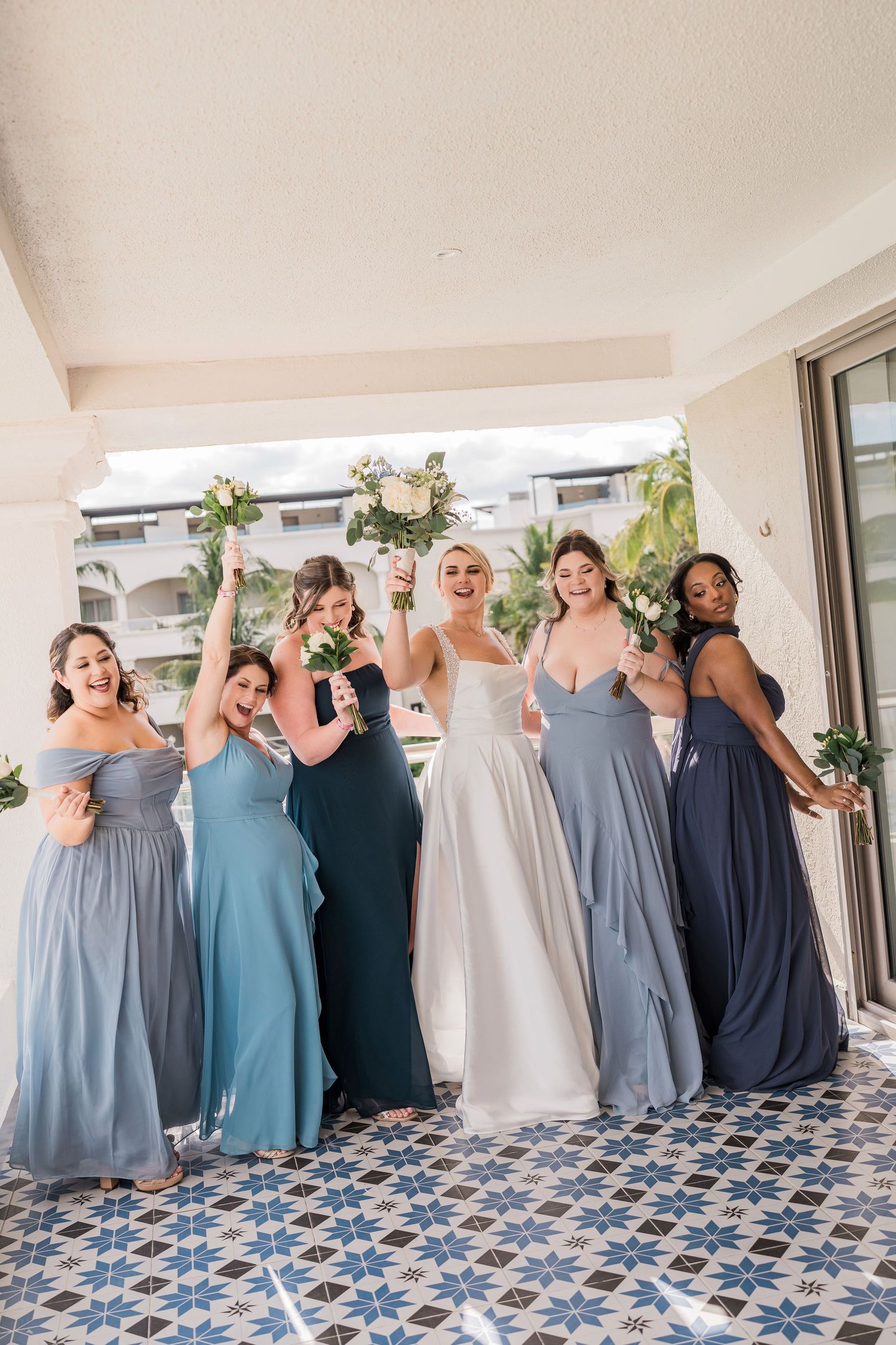Bride in white gown celebrates with five bridesmaids in blue dresses, raising bouquets on a patterned tile balcony.
