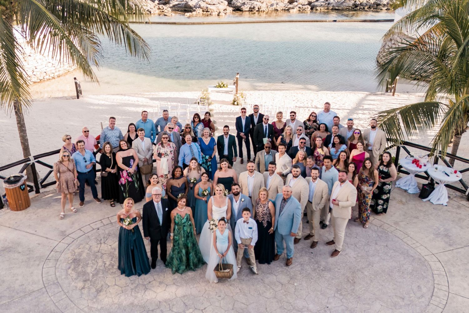 Large wedding group photo outdoors near a beach resort with palm trees and ocean views on a sunny day.