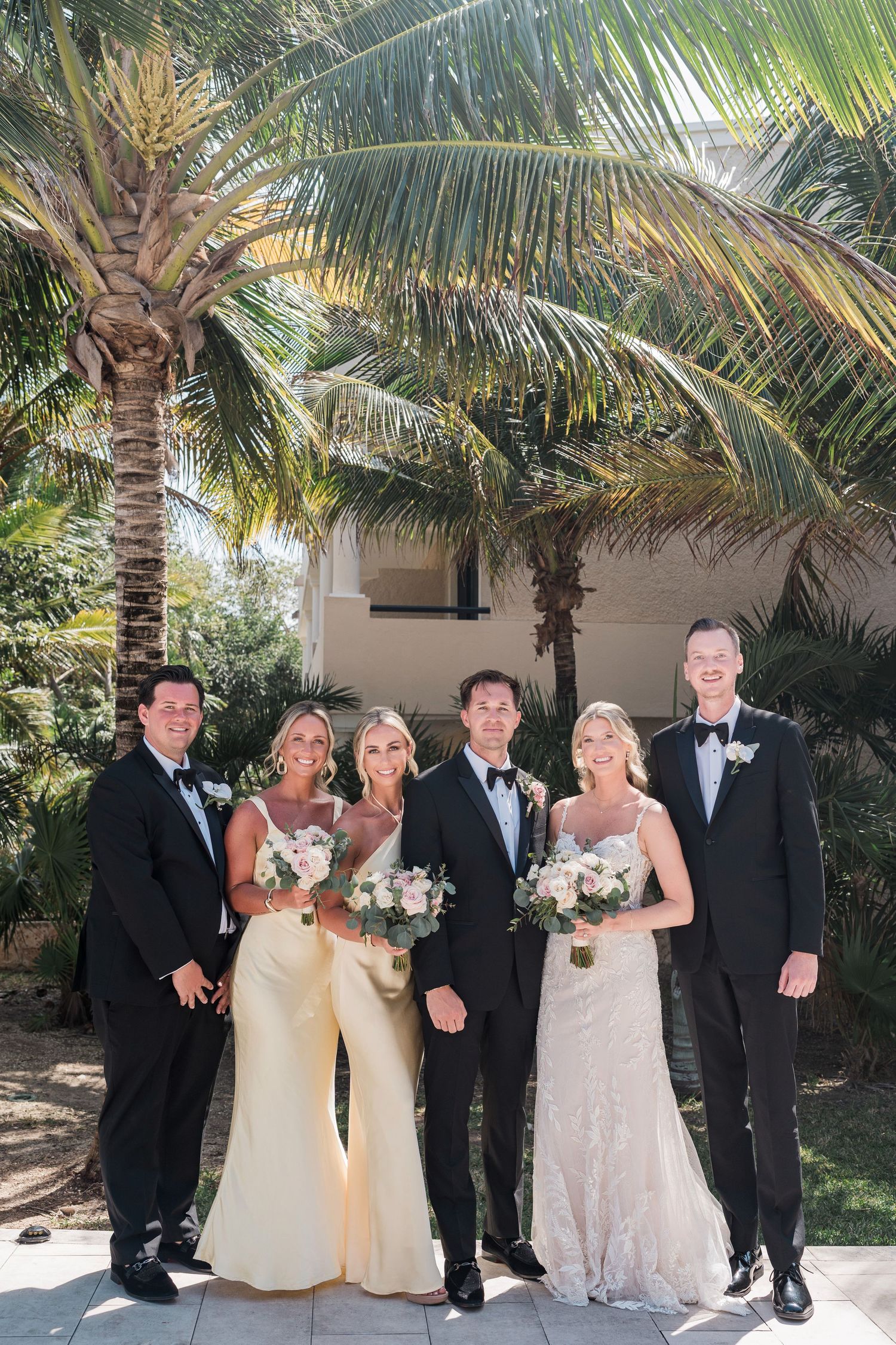 Wedding party posing outdoors near palm trees, bride in white gown, groom in tuxedo, guests in formal attire.