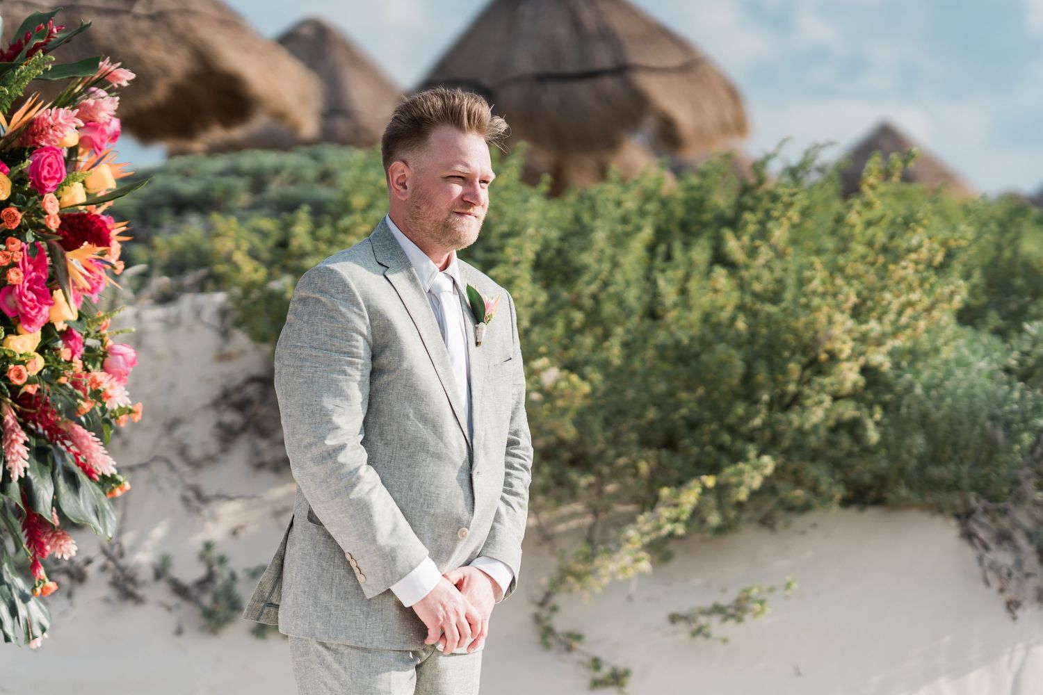 Groom in gray suit smiling at beach wedding ceremony with colorful floral arrangement and thatched huts in background.