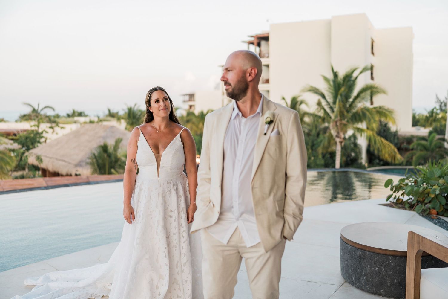 Bride in lace gown and groom in tan suit walk together near a resort pool with tropical palms at sunset.