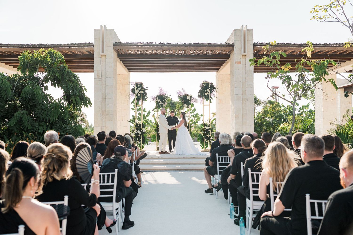 Elegant outdoor wedding ceremony with guests seated in white chairs along an aisle leading to couple under pergola.