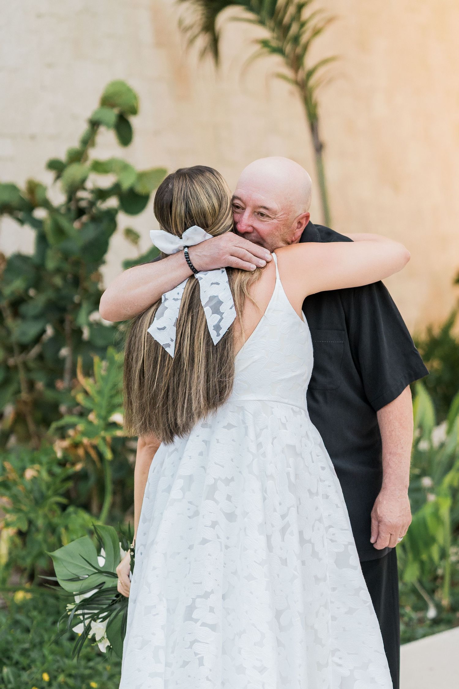 Bride in white lace dress hugging bald man in black suit at outdoor wedding ceremony near tropical plants.