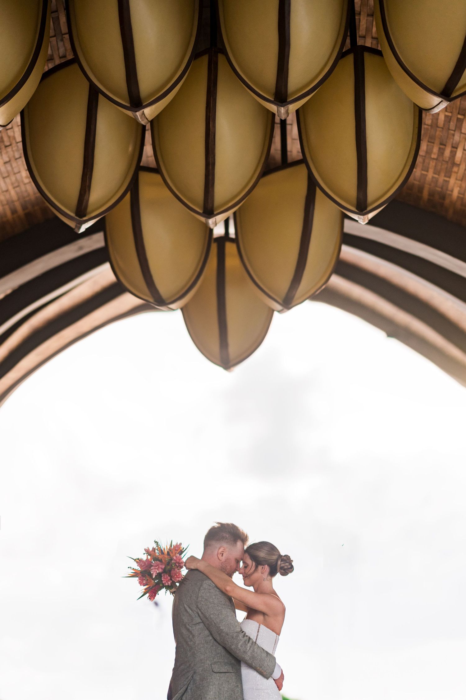 Bride and groom embrace under ornate chandelier through arched doorway, bride holding vibrant red bouquet.