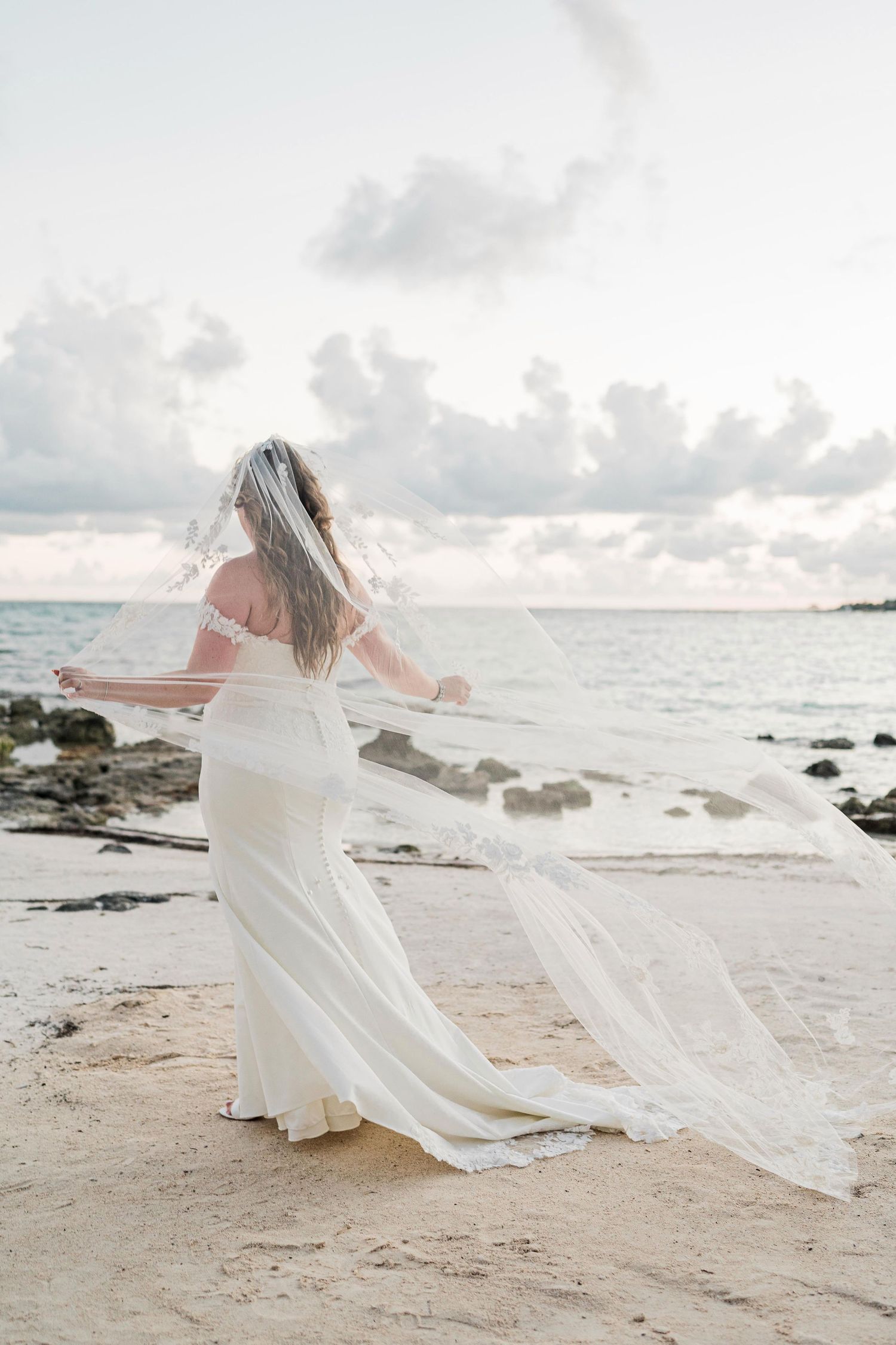 Bride in white gown and flowing veil stands on sandy beach with rocky shoreline and cloudy sky backdrop. Puerto Vallarta wedding photographe