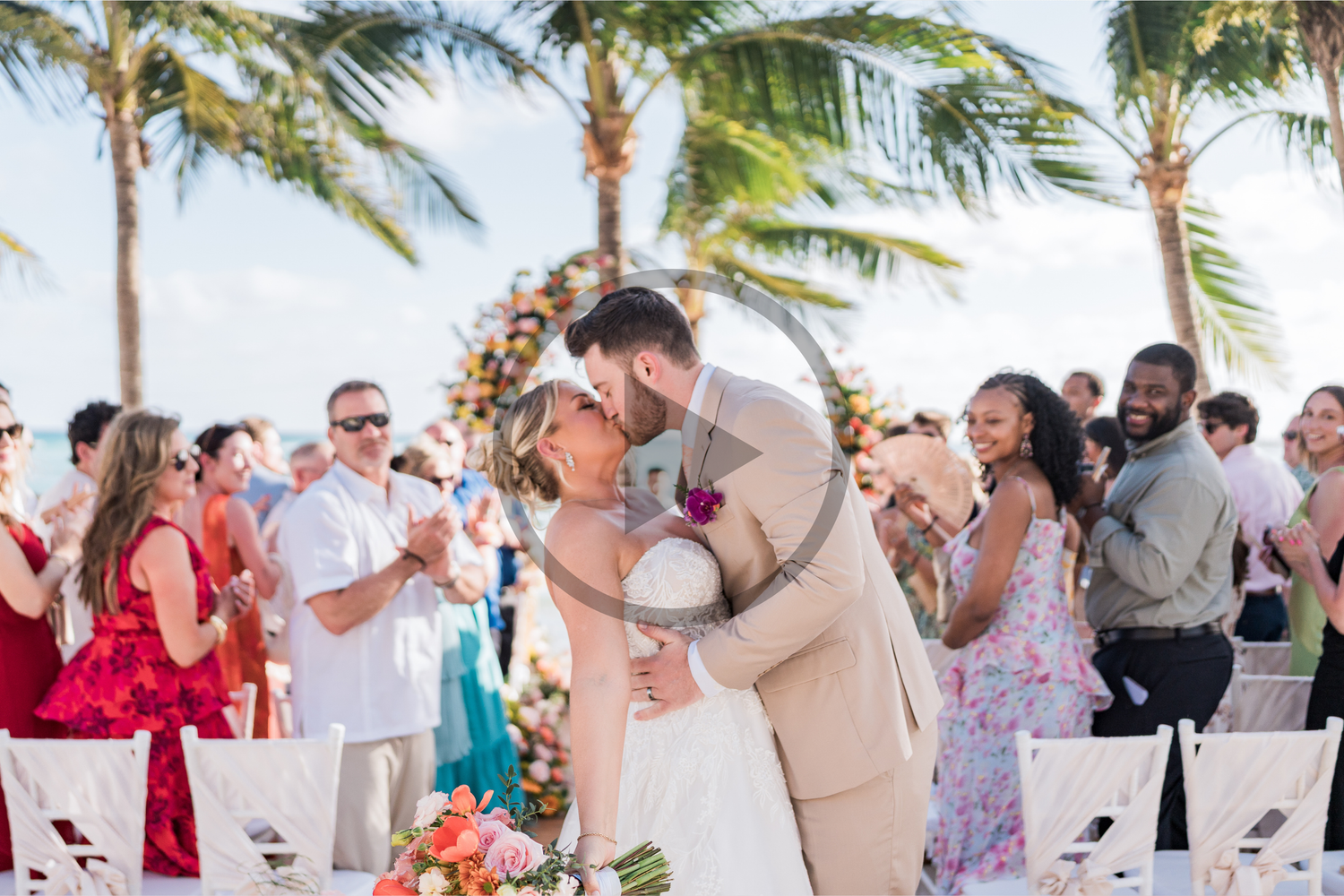 Bride and groom share first kiss at tropical beach wedding ceremony with palm trees and guests celebrating at Barceló Maya Palace Resort