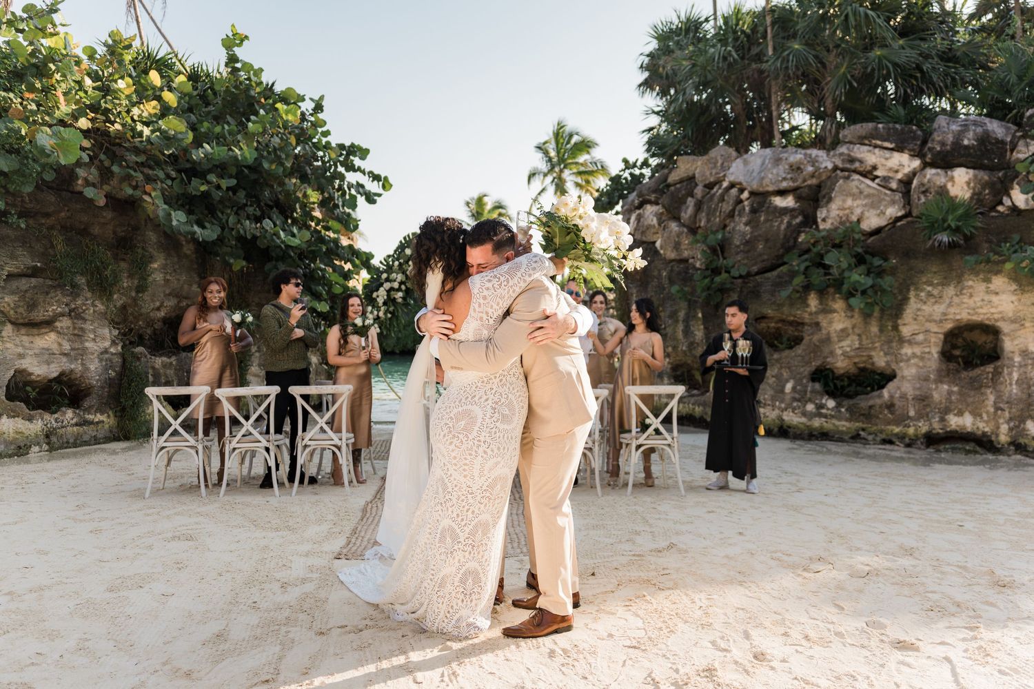 Xcaret Arte Wedding - Couple shares first dance embrace on sandy beach wedding venue with guests and tropical rock backdrop.