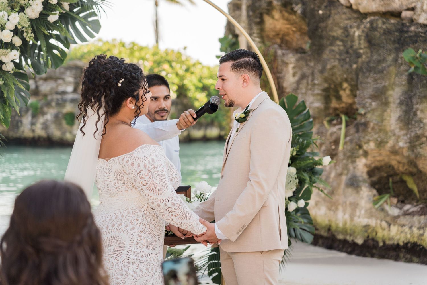 Xcaret Arte Wedding - Couple exchanging vows at a tropical beach wedding ceremony with lush greenery and a rocky backdrop.
