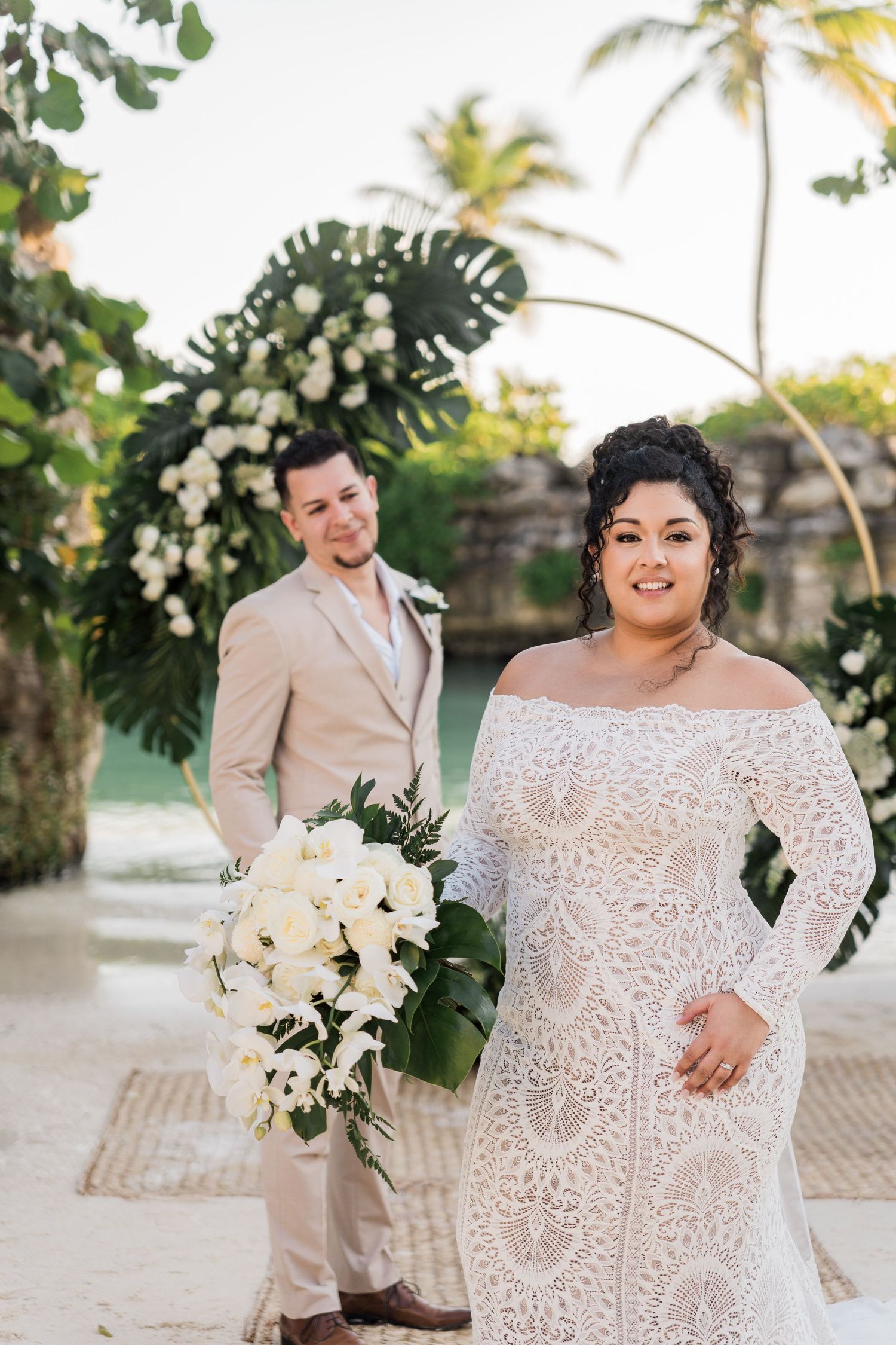Xcaret Arte Wedding - Bride in lace off-shoulder gown holds white bouquet as groom in tan suit stands before floral arch at beach wedding.