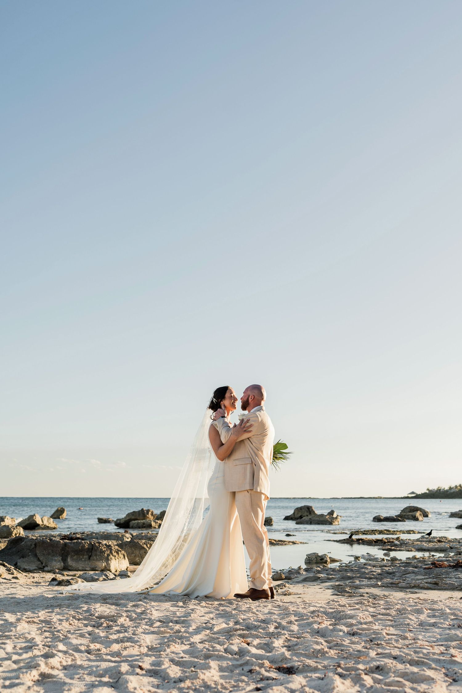 Bride and groom embrace on sandy beach at sunset, bride in flowing white gown with veil, groom in beige suit.