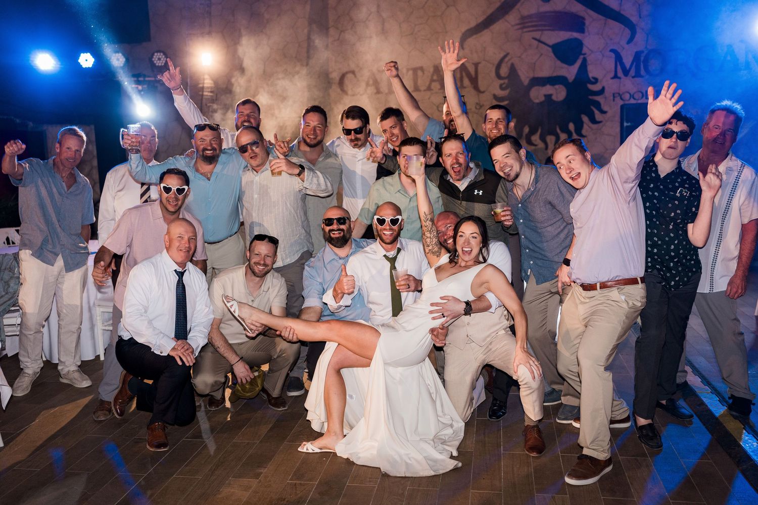 Large wedding party group photo with bride in white dress being lifted by groomsmen on a dance floor.