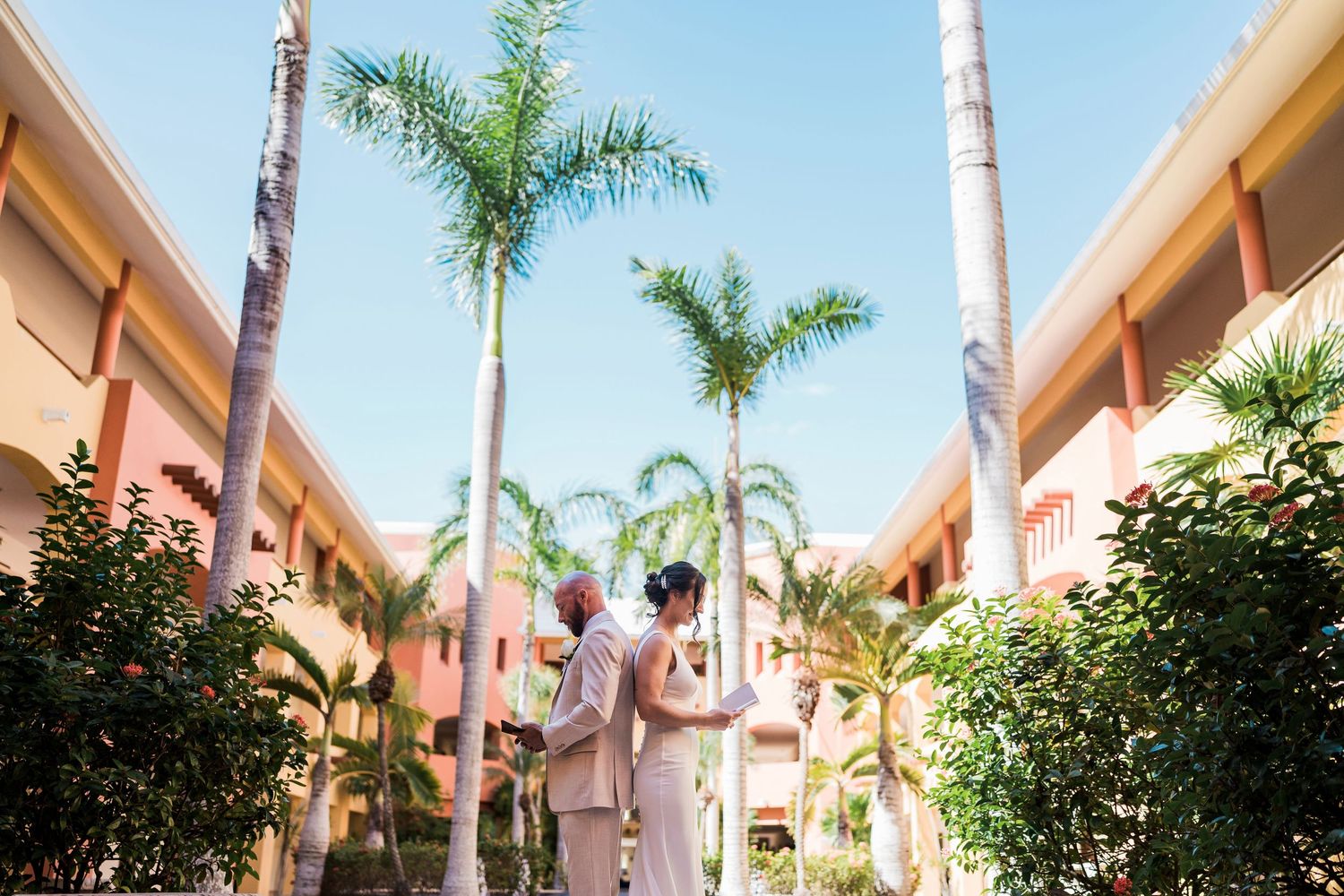 Couple in formal attire stands among tall palm trees at a tropical resort on their wedding day.