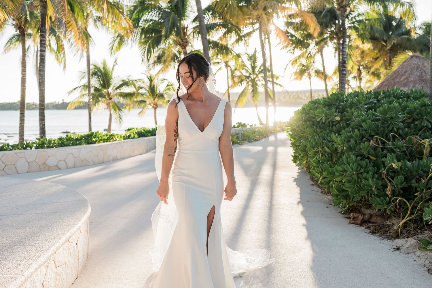 Bride in elegant white gown walks along tropical beach path lined with palm trees at golden hour.
