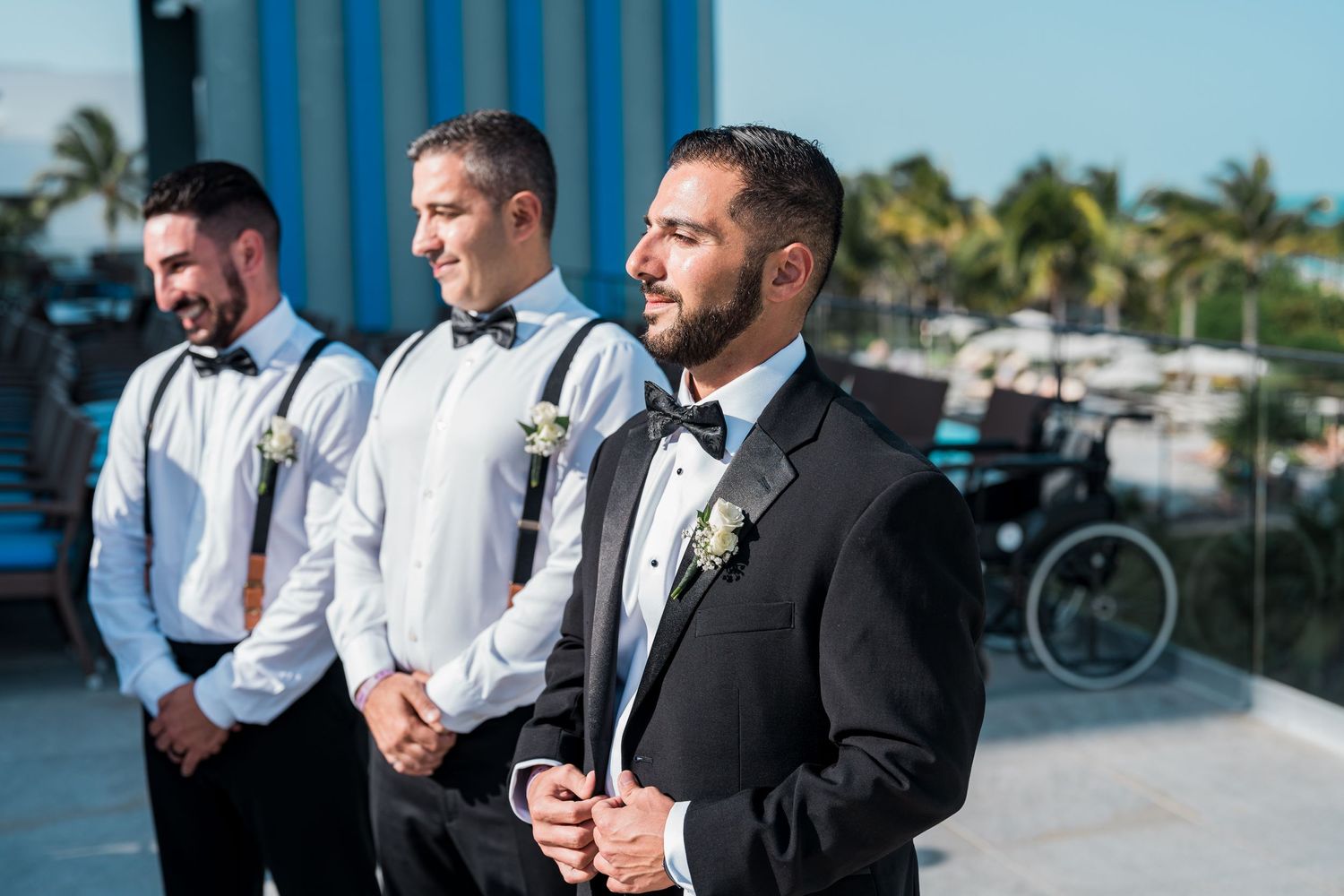 Groom and groomsmen in black tuxedos with bow ties and suspenders await ceremony at oceanside wedding venue.
