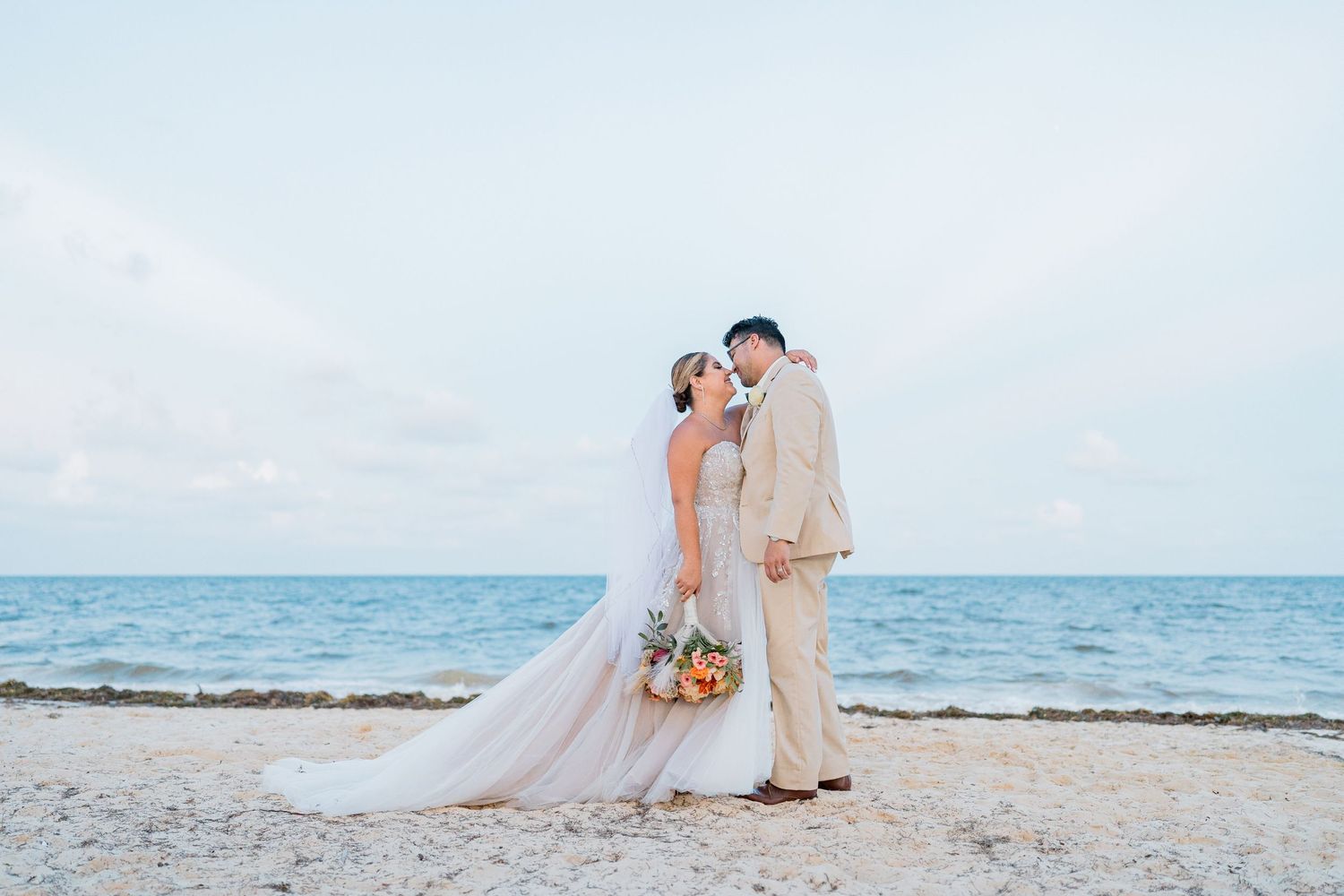 Bride and groom standing close together on sandy beach with turquoise ocean and cloudy sky behind them.
