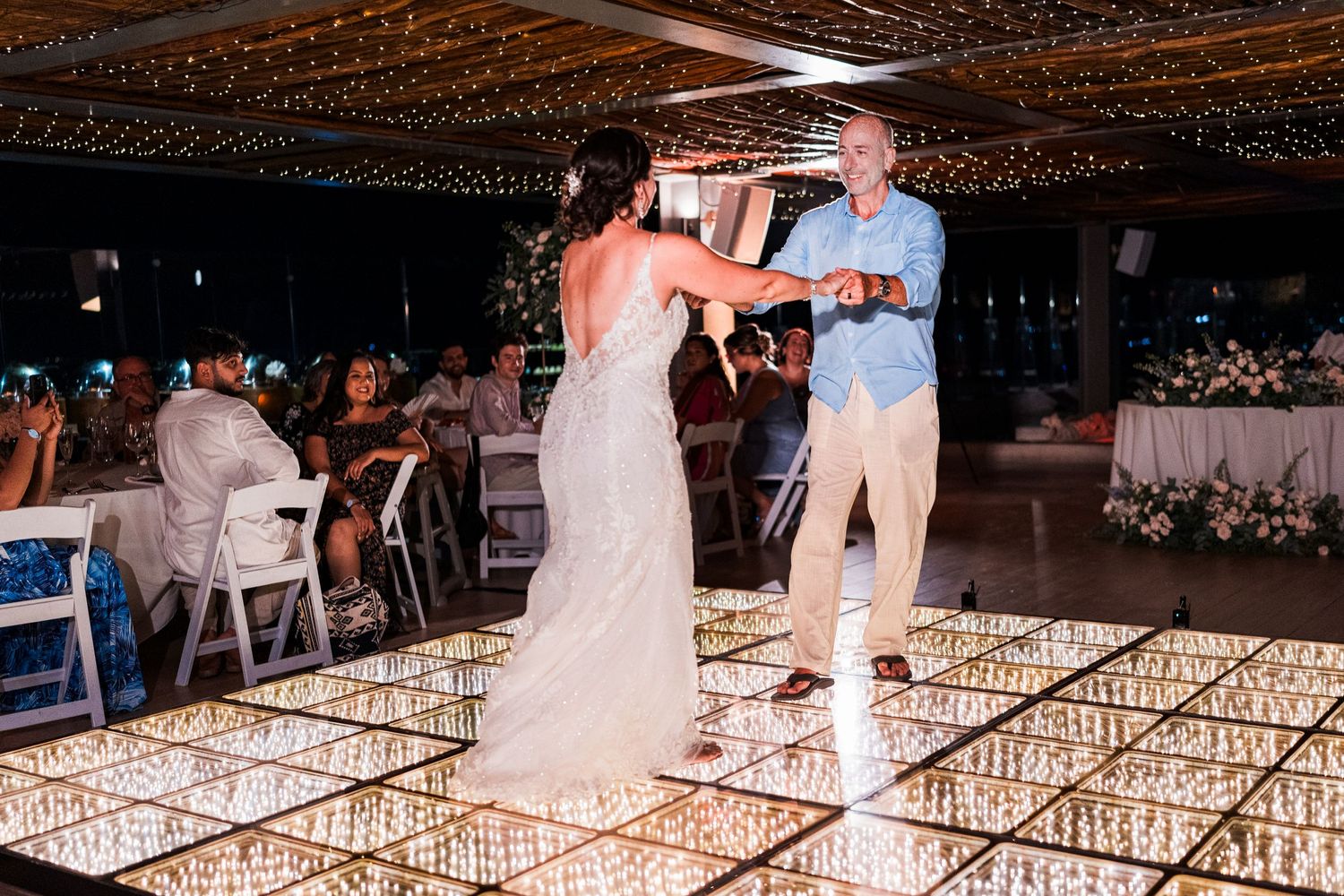 Bride dances with her father on a glowing mirrored dance floor at a beach wedding reception.