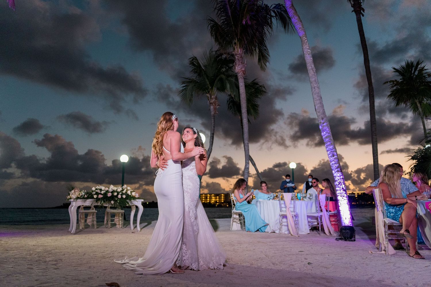 Two brides share first dance on sandy beach at sunset with guests seated nearby.