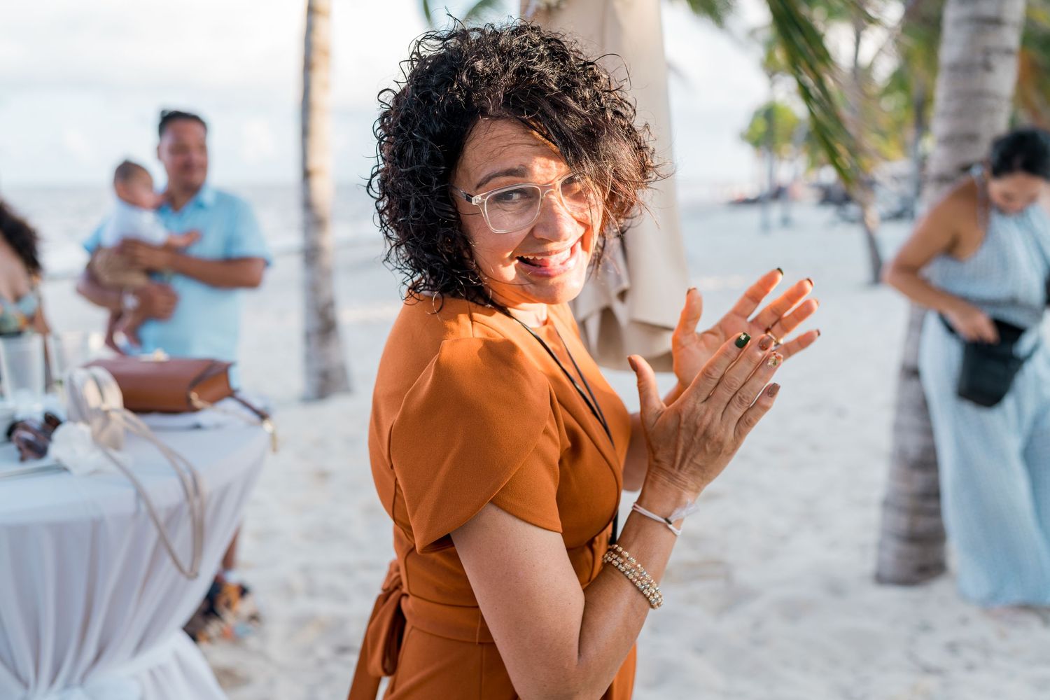 Smiling woman in orange dress claps her hands at a tropical beach wedding reception near palm trees.