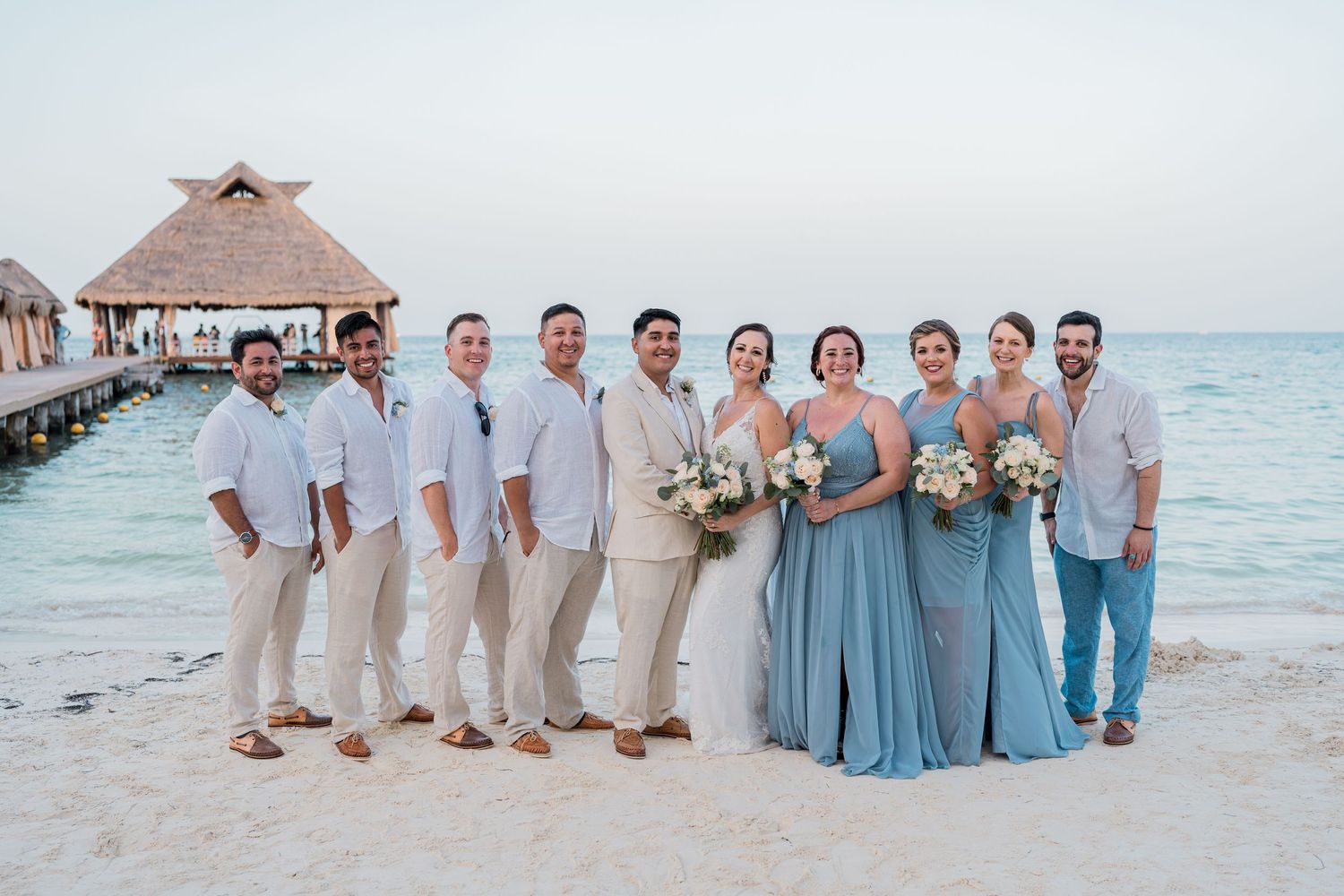 Wedding party poses on sandy beach in blue and beige attire with overwater bungalow backdrop.
