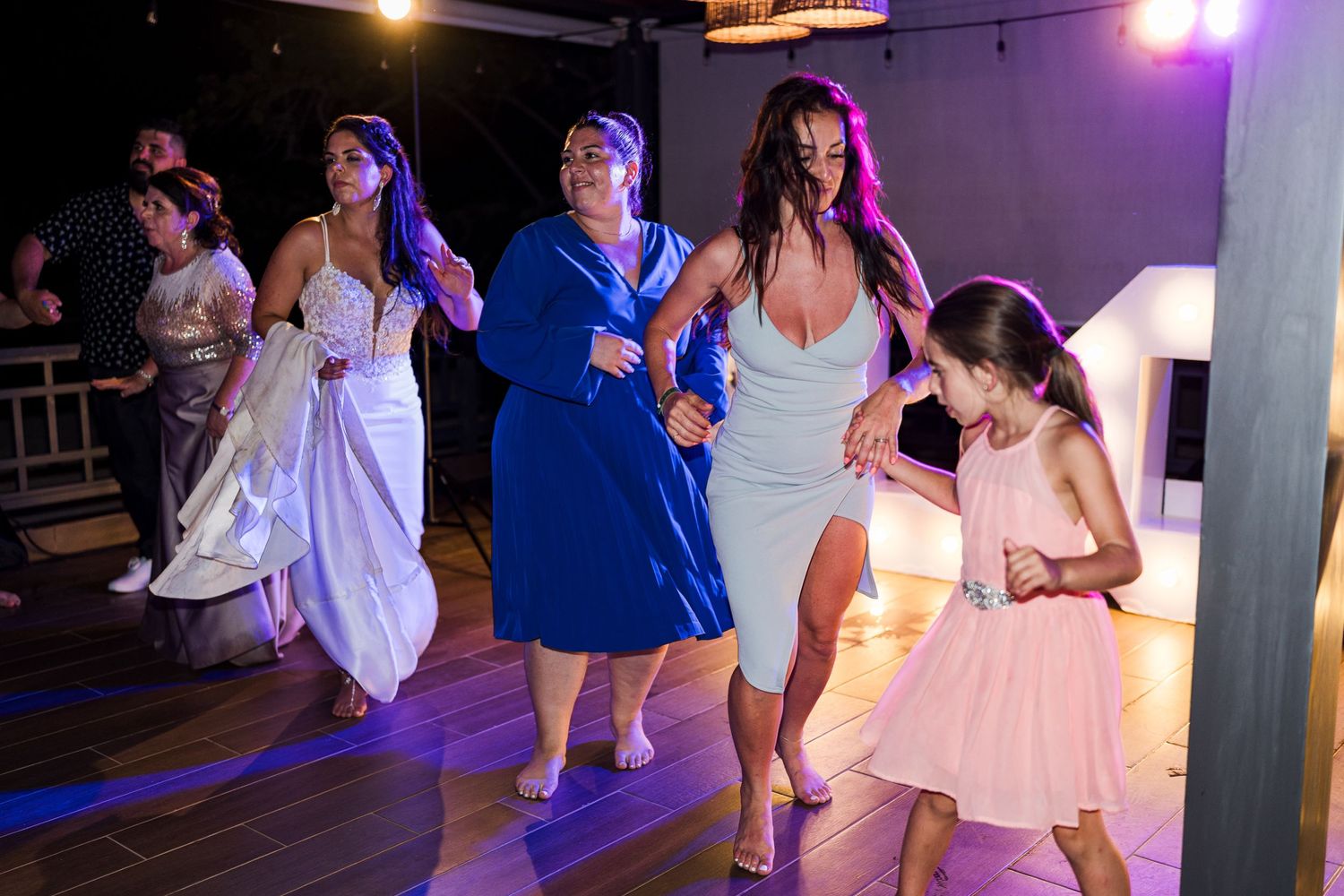 Group of women and a young girl dance together energetically at a beach wedding reception.