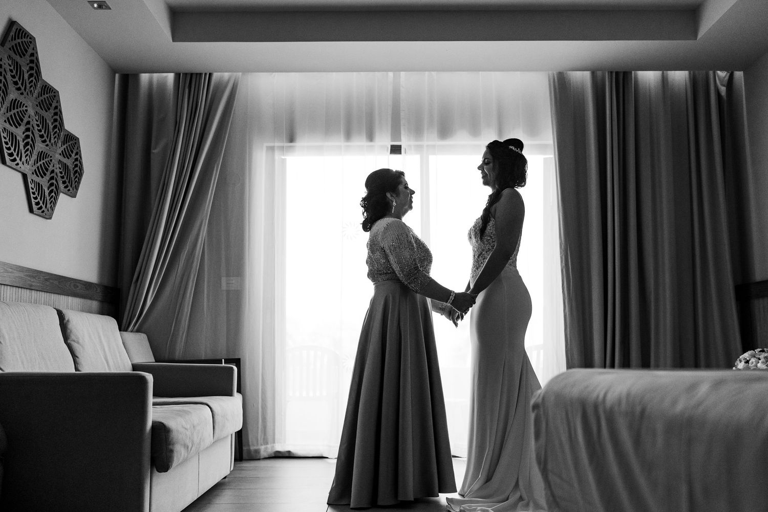 Black and white silhouette of bride and bridesmaid holding hands by window in hotel room.