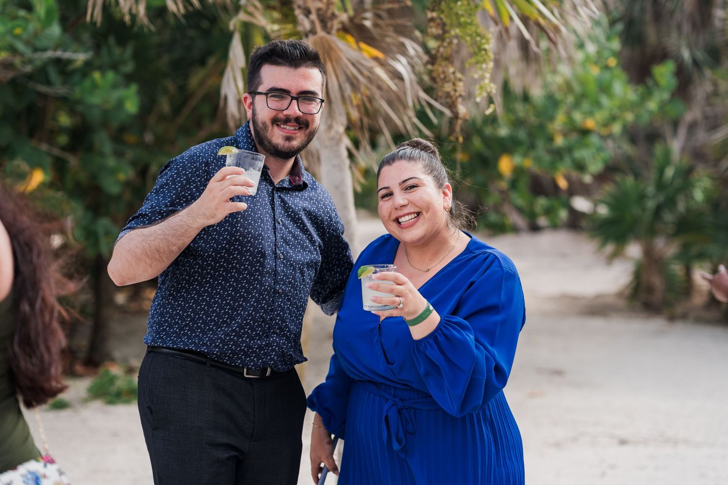 Man and woman smiling and holding drinks at an outdoor beach wedding reception.