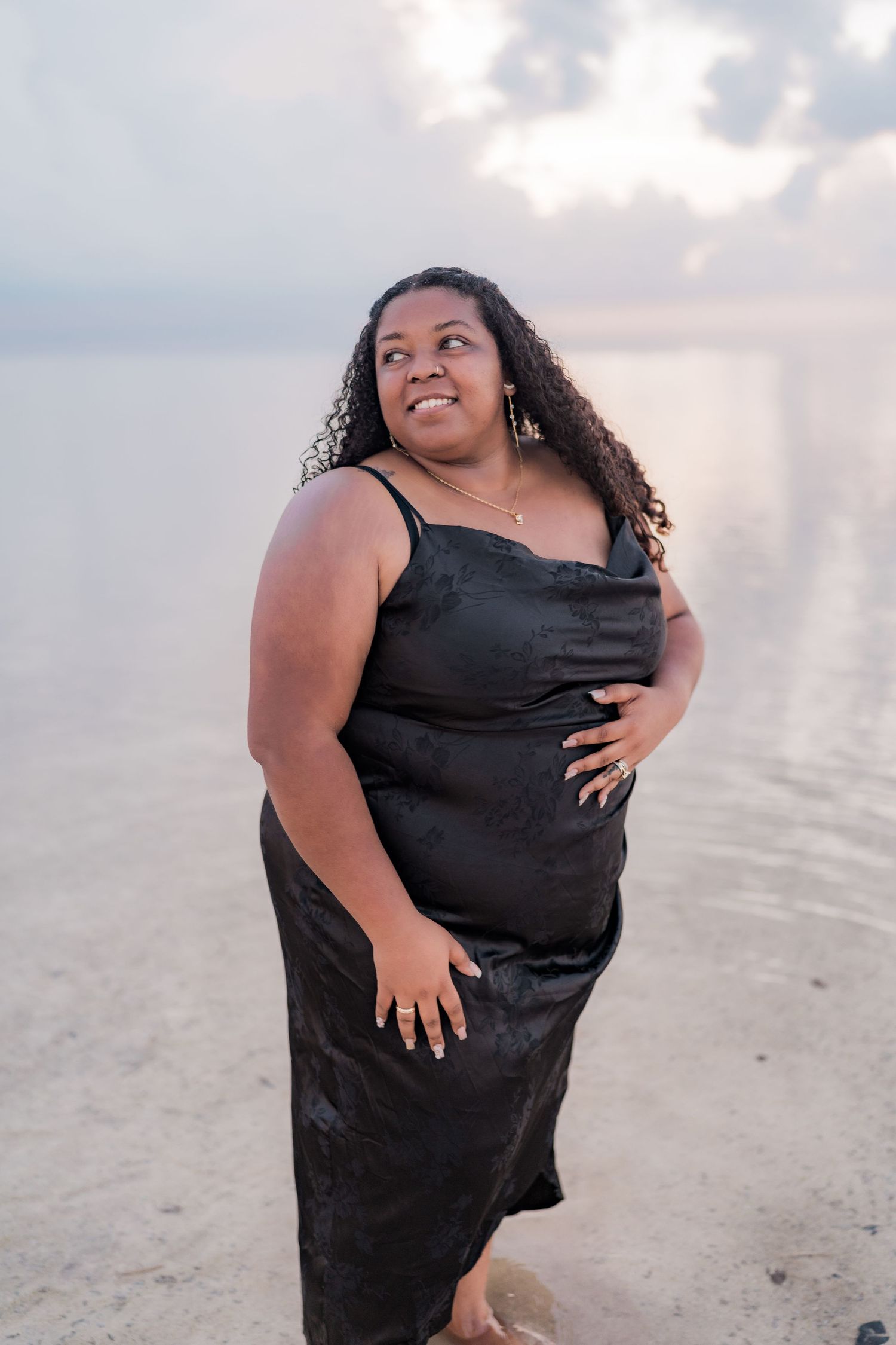 Woman in black satin dress posing confidently on a beach at sunset.