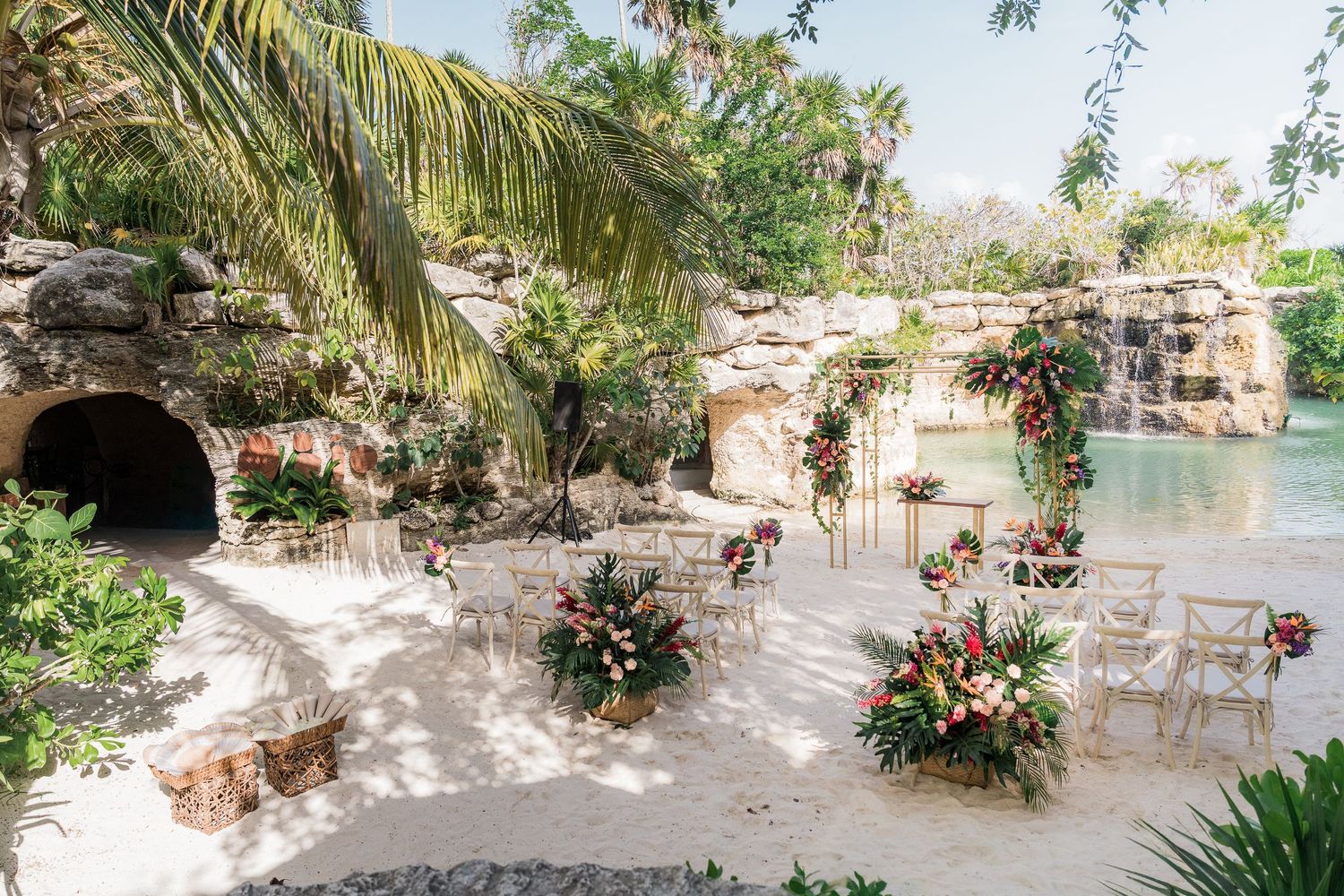 Hotel Xcaret México Wedding - Tropical outdoor wedding ceremony setup on sandy beach with floral arches, by a lagoon.