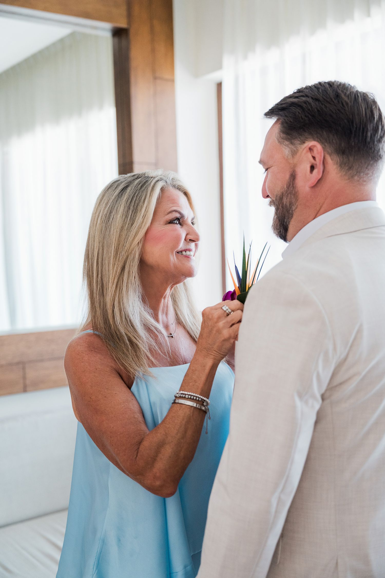A smiling blonde woman in a blue dress pins a boutonniere on a man in a white suit jacket.
