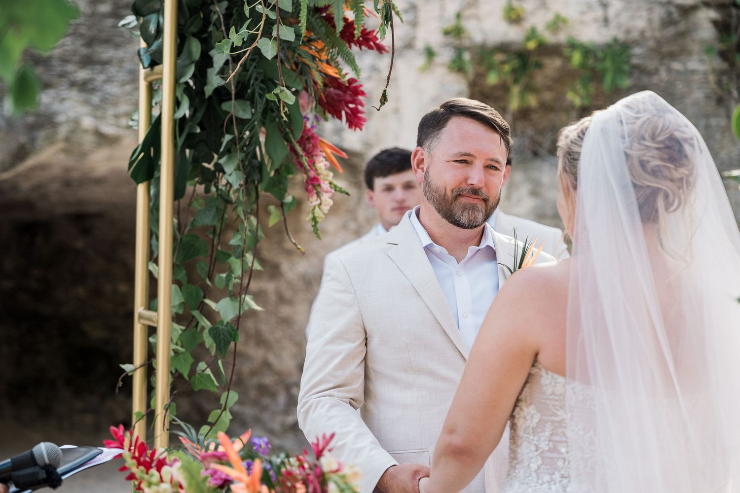 Groom in beige suit smiles at bride during outdoor wedding ceremony with colorful floral arch backdrop.