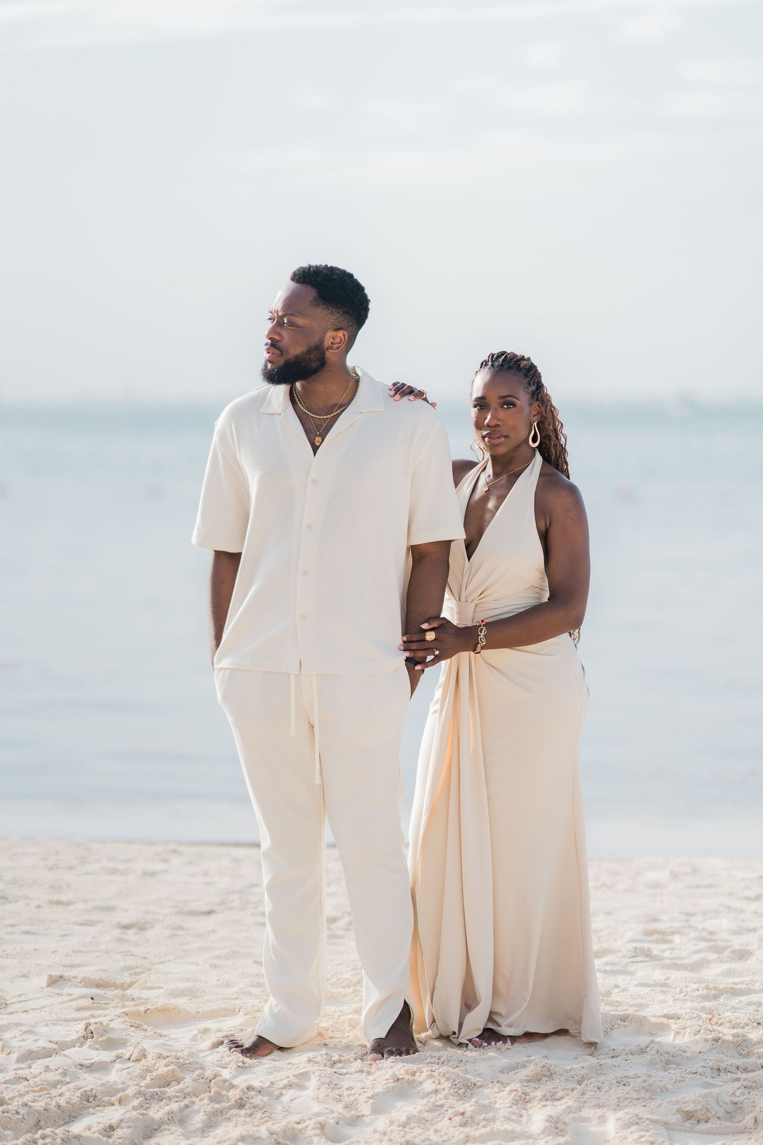 Black couple in matching cream outfits posing together on a sandy beach.