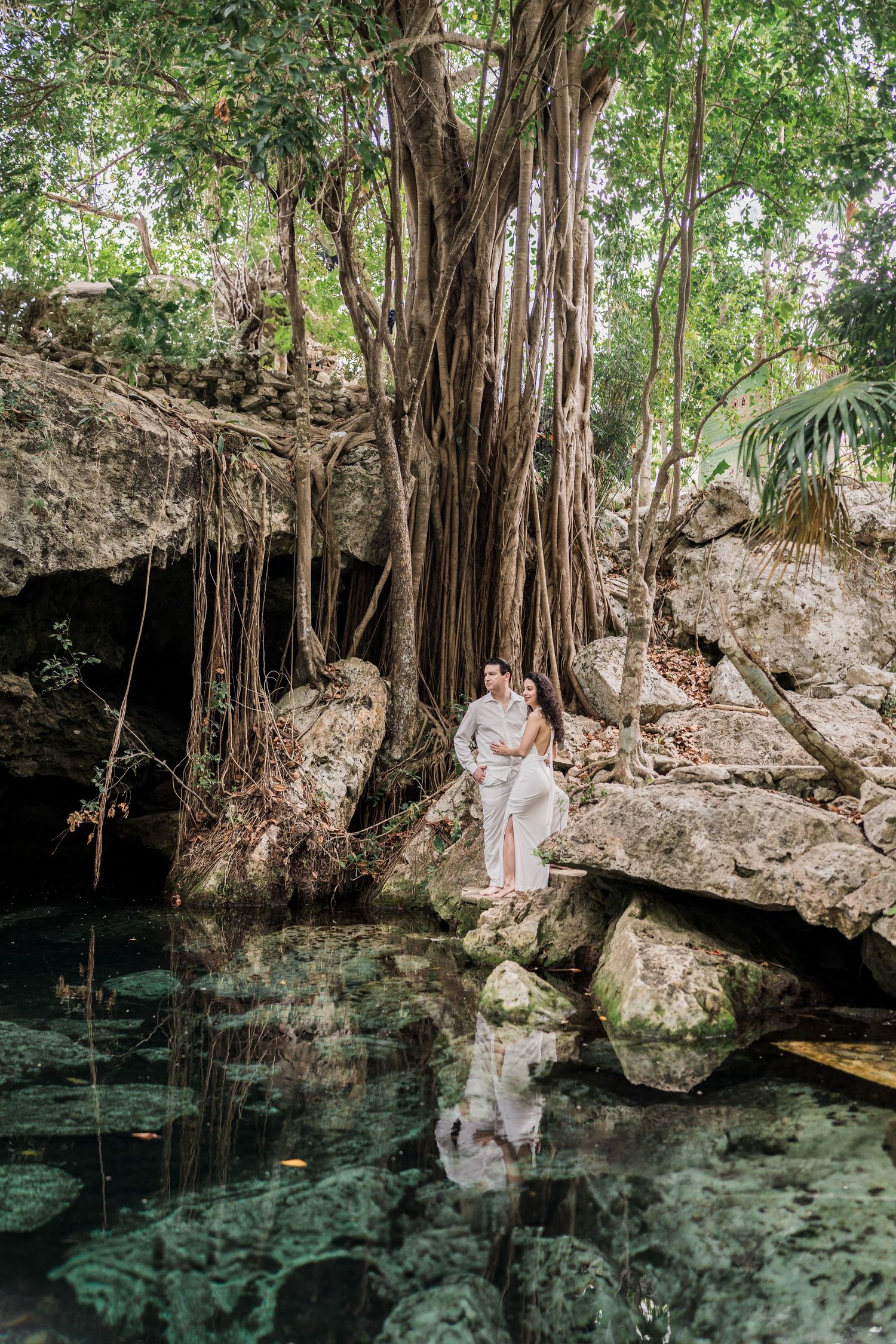 Couple embracing on rocks beside a crystal-clear cenote surrounded by lush tropical trees and hanging roots in Mexico.