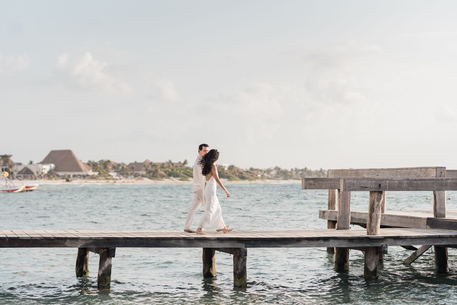 A couple shares a romantic embrace on a wooden pier overlooking turquoise ocean waters at a tropical resort.