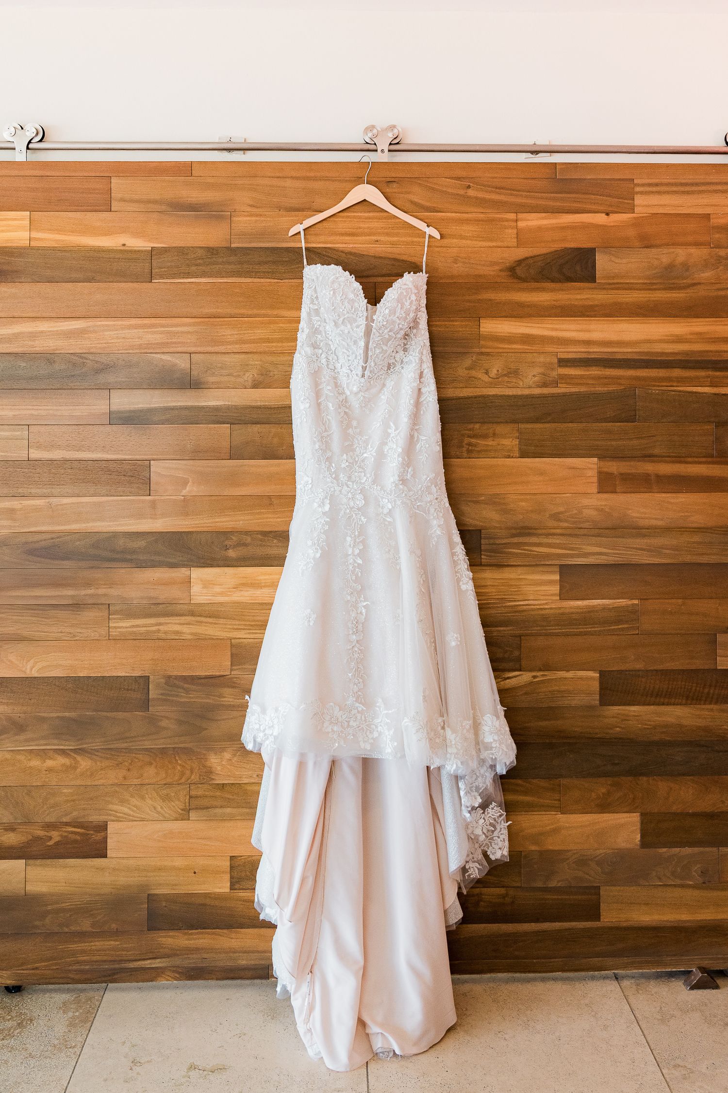 Elegant strapless lace wedding dress with blush skirt hanging on wooden wall.