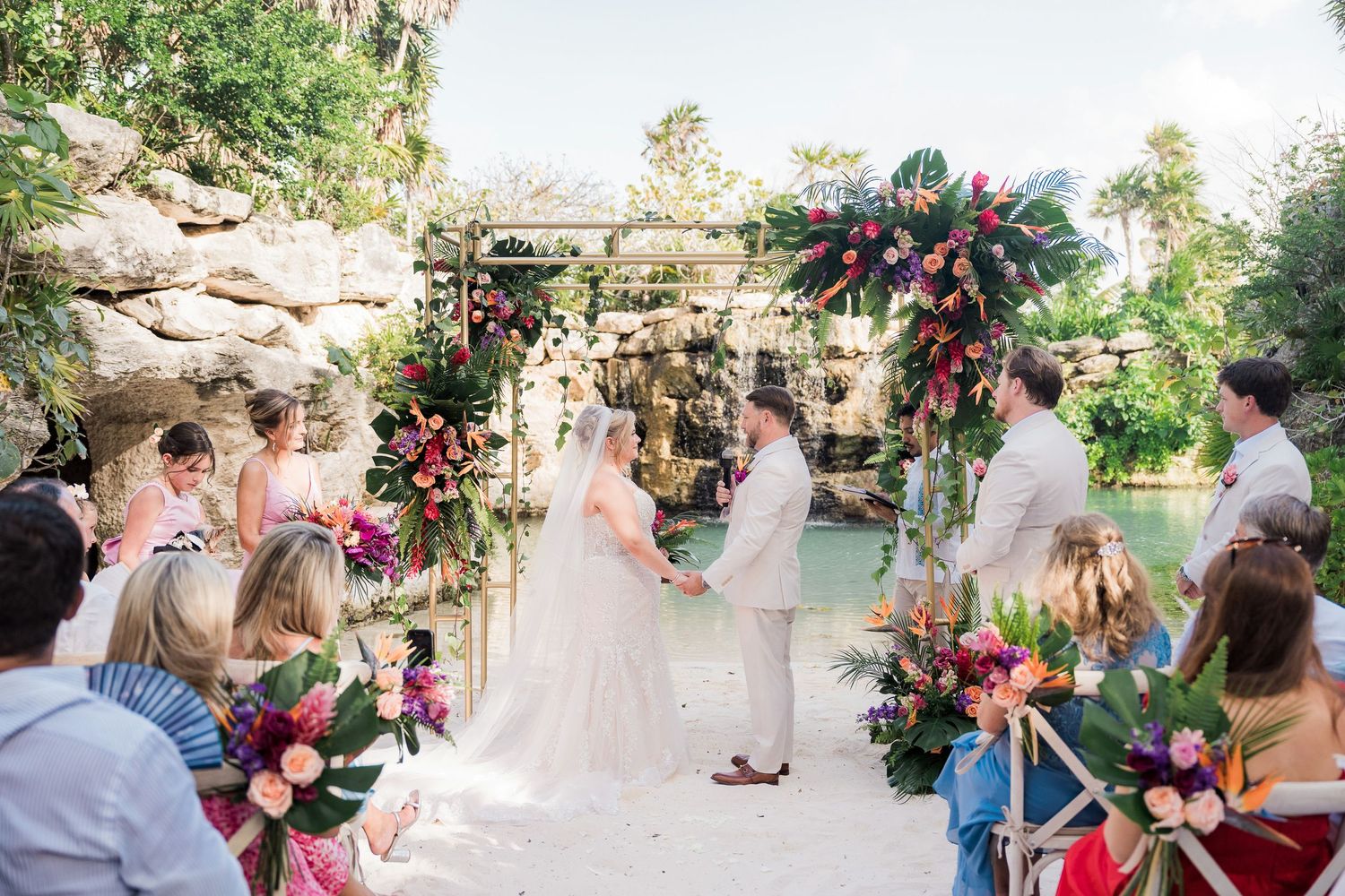 Colorful tropical wedding ceremony outdoors with floral arch, bride and groom holding hands, guests seated along sandy aisle.